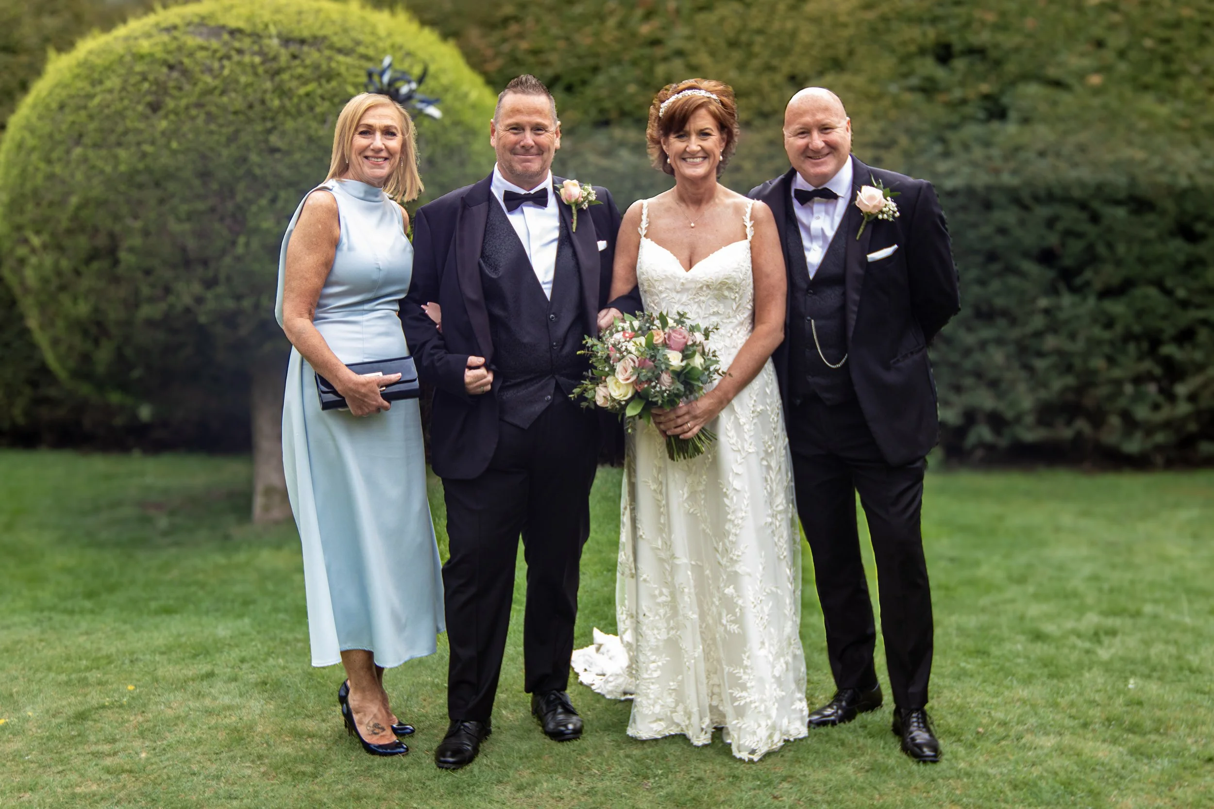 Group of four people dressed in formal wedding attire, standing on a lawn with trees in the background. The woman in the white wedding dress holds a bouquet of flowers. The men are in black tuxedos with bow ties, and the woman on the left is in a light blue dress.