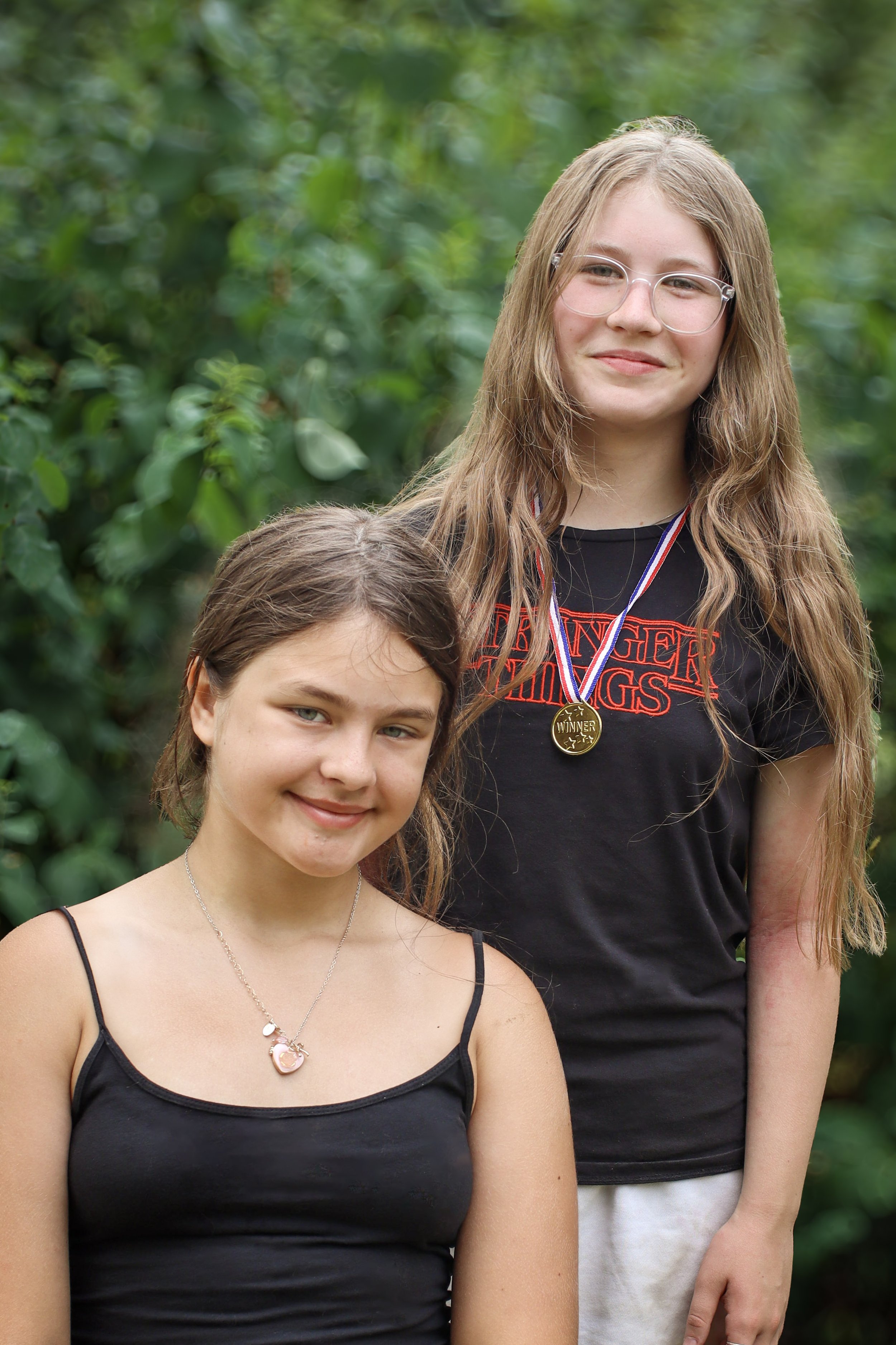 Two young girls standing outdoors with greenery in the background, smiling at the camera. One girl is wearing a black spaghetti strap top and a necklace, and the other girl is wearing glasses, a black T-shirt with red lettering, and a medal around her neck.