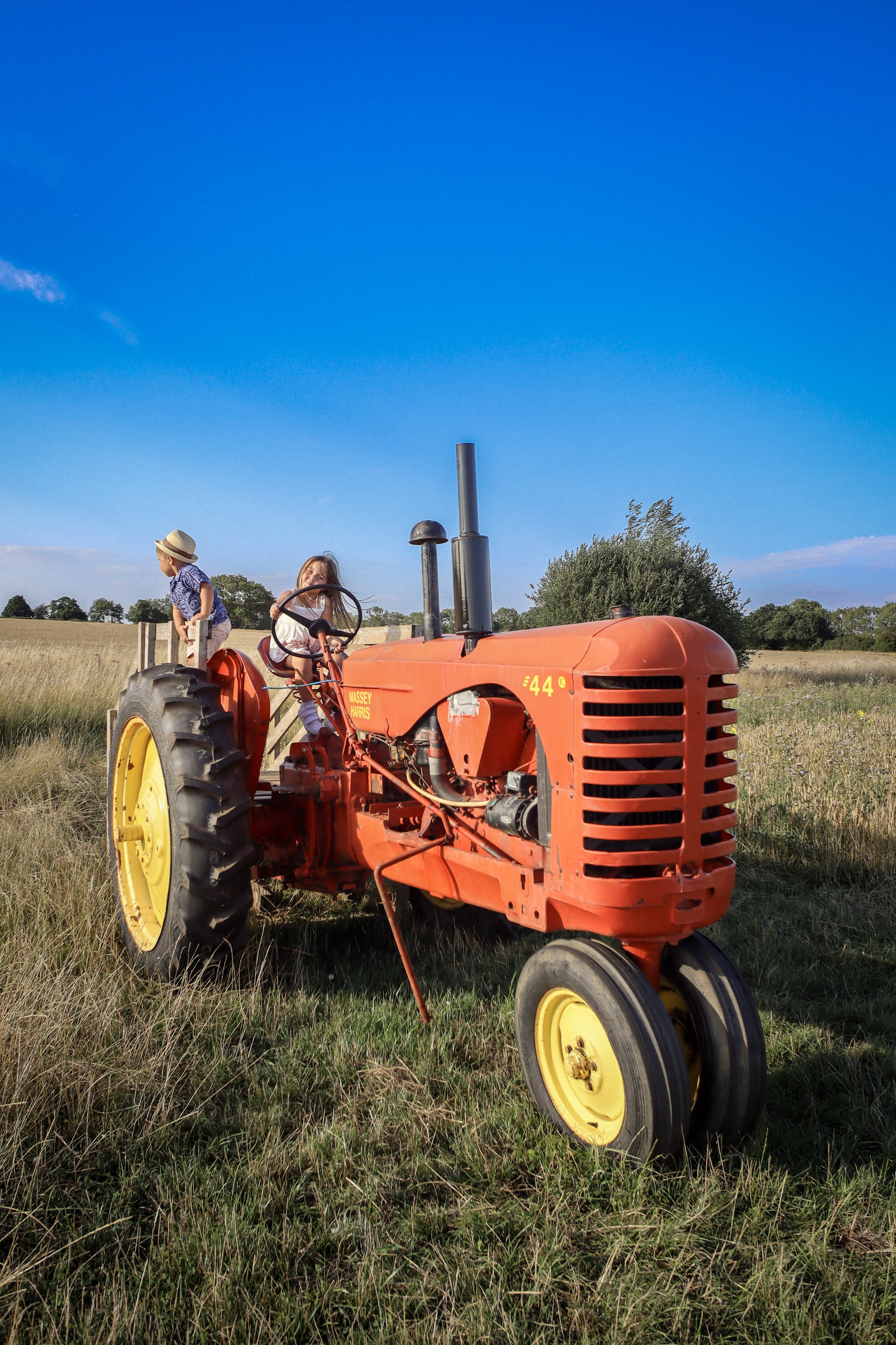 Two children sitting on an orange vintage tractor in a grassy field under a bright blue sky.