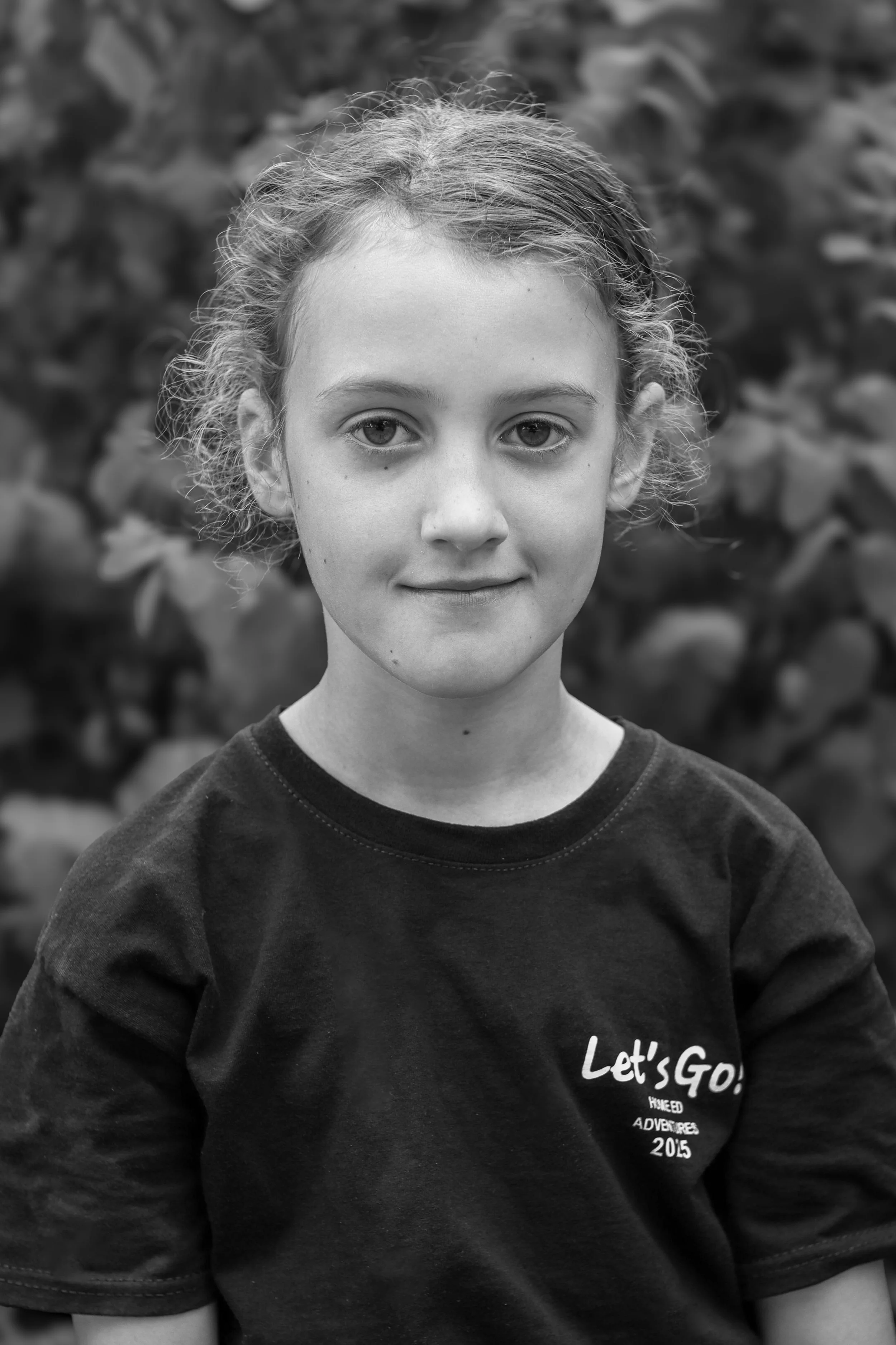 A young girl with curly hair and light skin, wearing a dark shirt with the text 'Let's Go' on it, standing outdoors with a blurred natural background.
