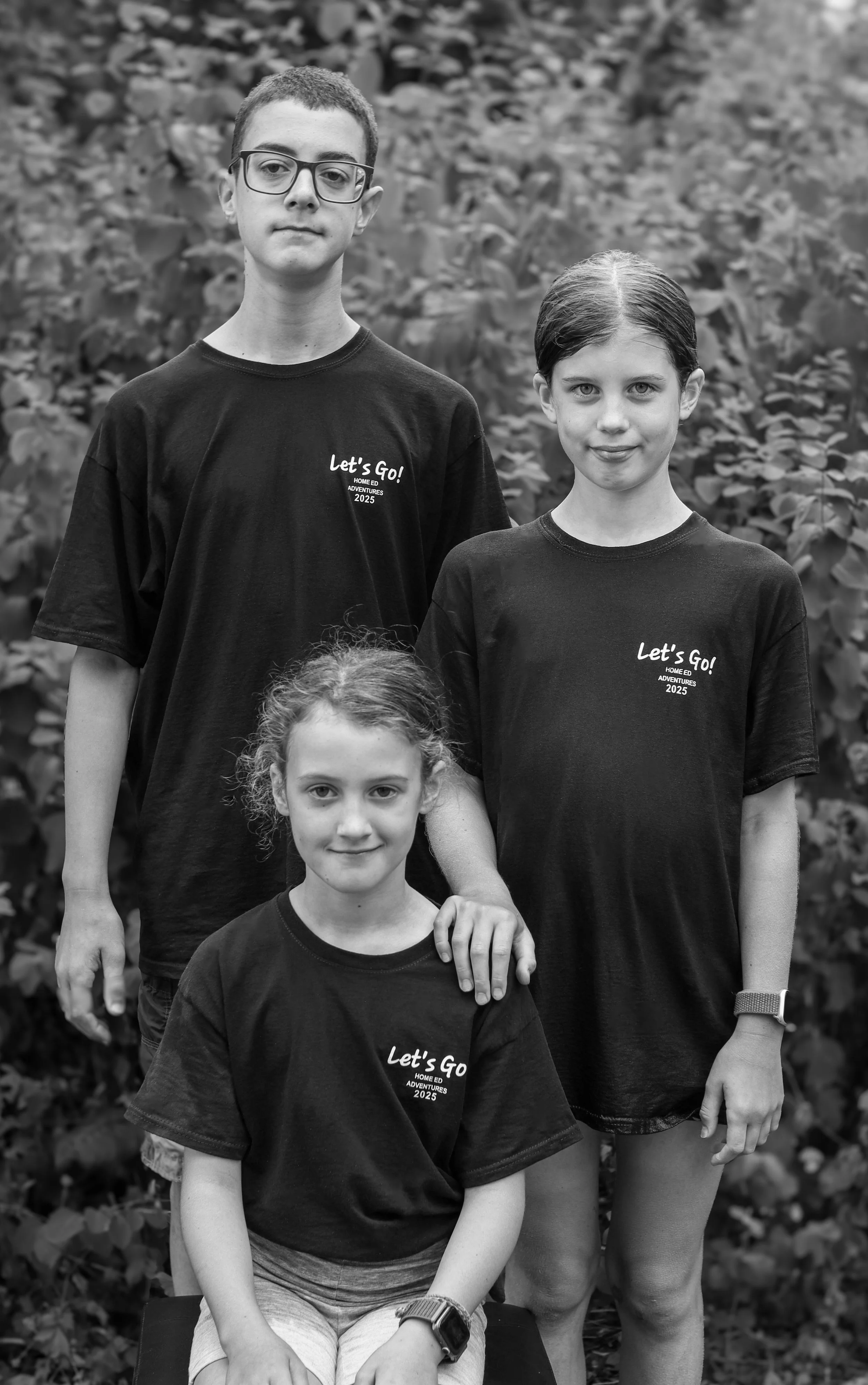 Black and white photo of three children, two girls and one boy, standing outdoors in front of a leafy background. They are wearing matching T-shirts that read 'Let's Go! Home Ed Adventures 2025'. The girl in the front is sitting, while the girl on the right and the boy are standing behind her.