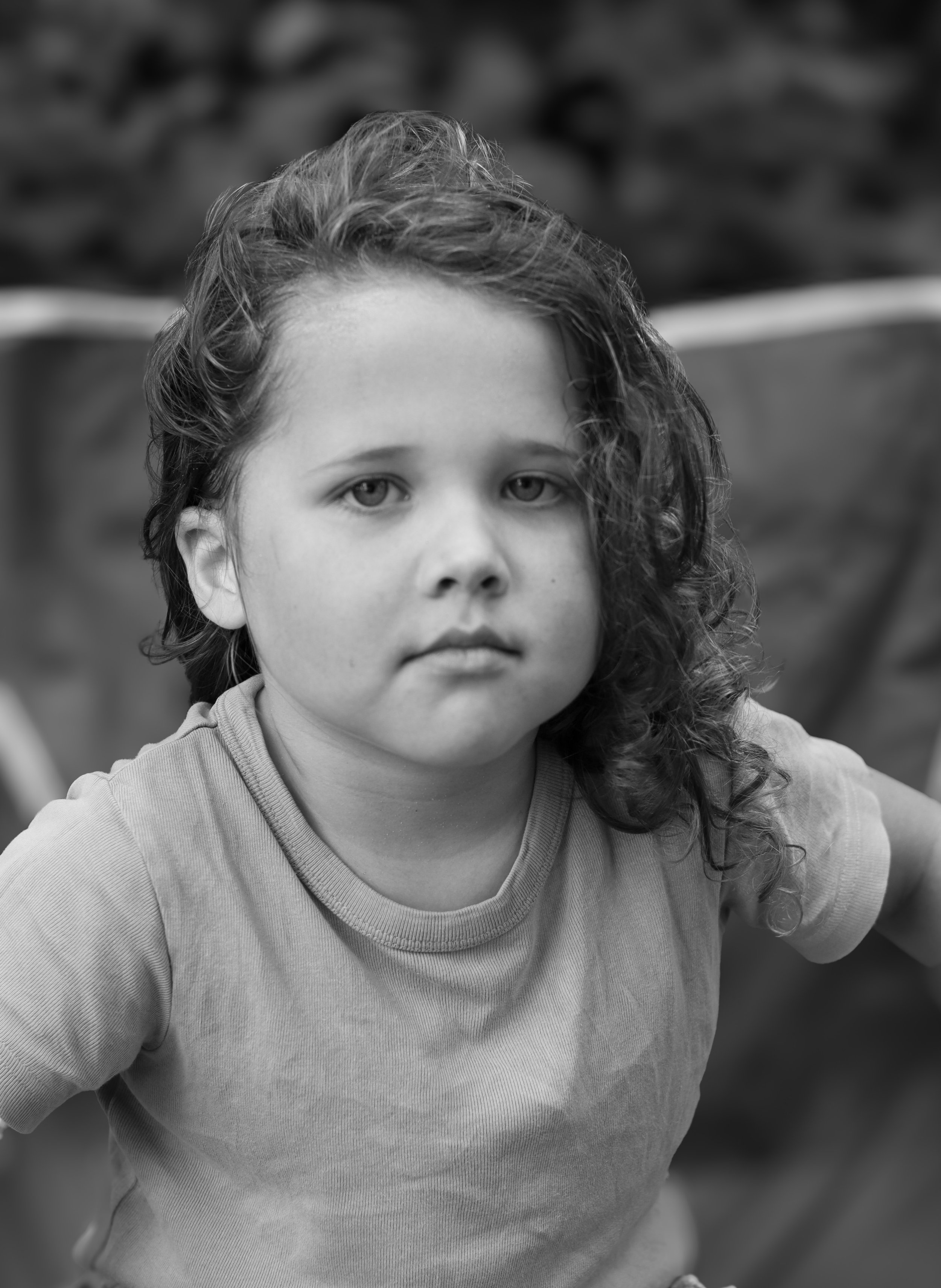 A young girl with curly hair and a serious expression on her face, wearing a plain T-shirt, outdoors with blurred natural background.