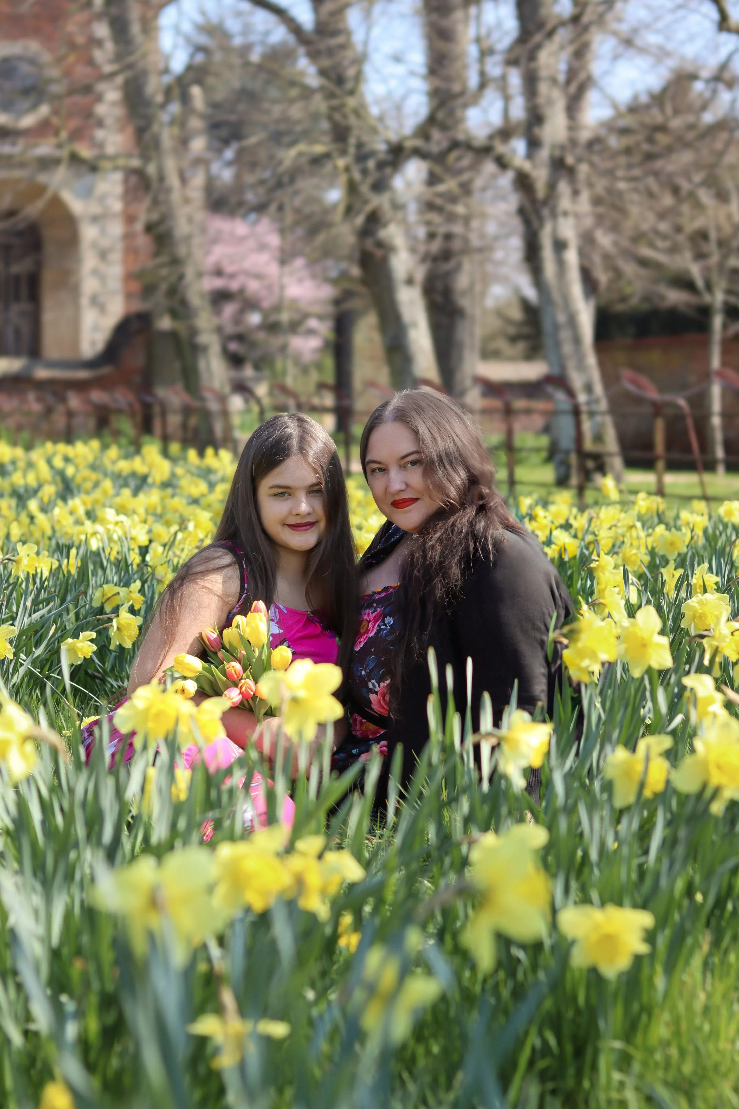 A woman and a girl sitting in a field of yellow flowers, holding a bouquet of similar flowers, with trees and a building in the background.