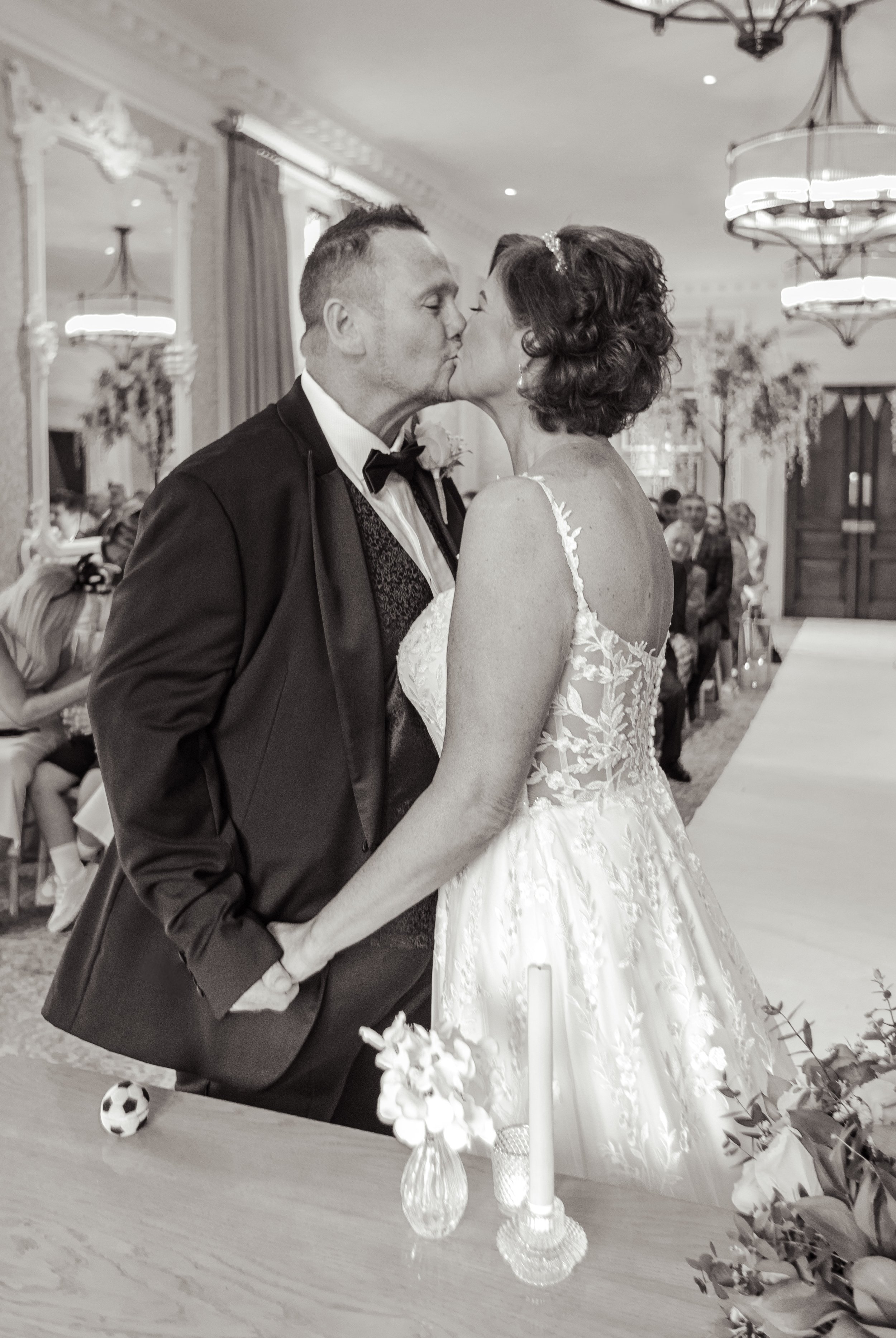 A black-and-white photo of a bride and groom kissing during their wedding ceremony. The bride is wearing a lace wedding gown and the groom is dressed in a tuxedo. They are holding hands, and the background shows wedding guests seated in a decorated v