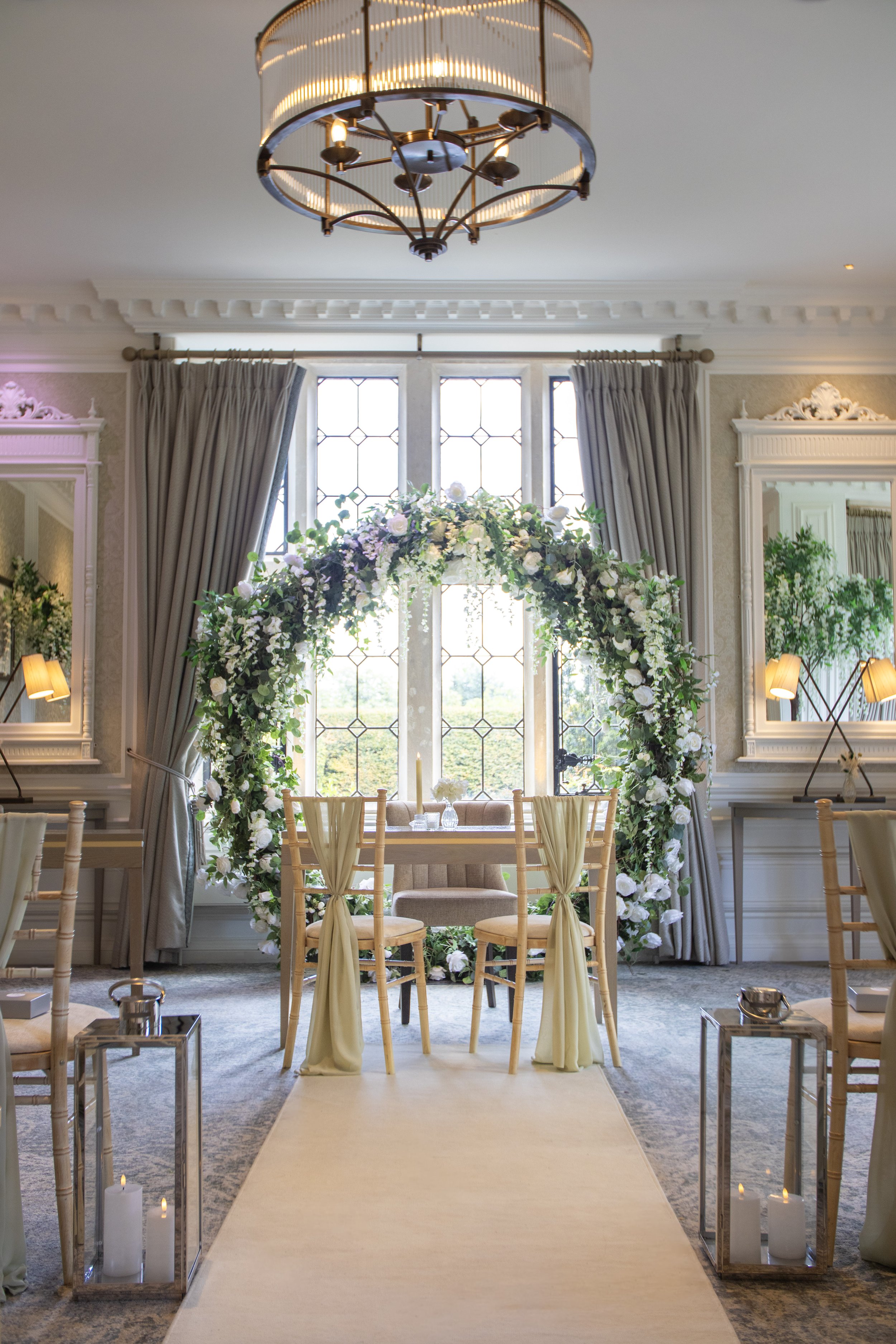 A decorated wedding altar with an arch of white and purple flowers, set in front of large stained glass windows, in a brightly lit room with gray curtains and mirrors on the walls.