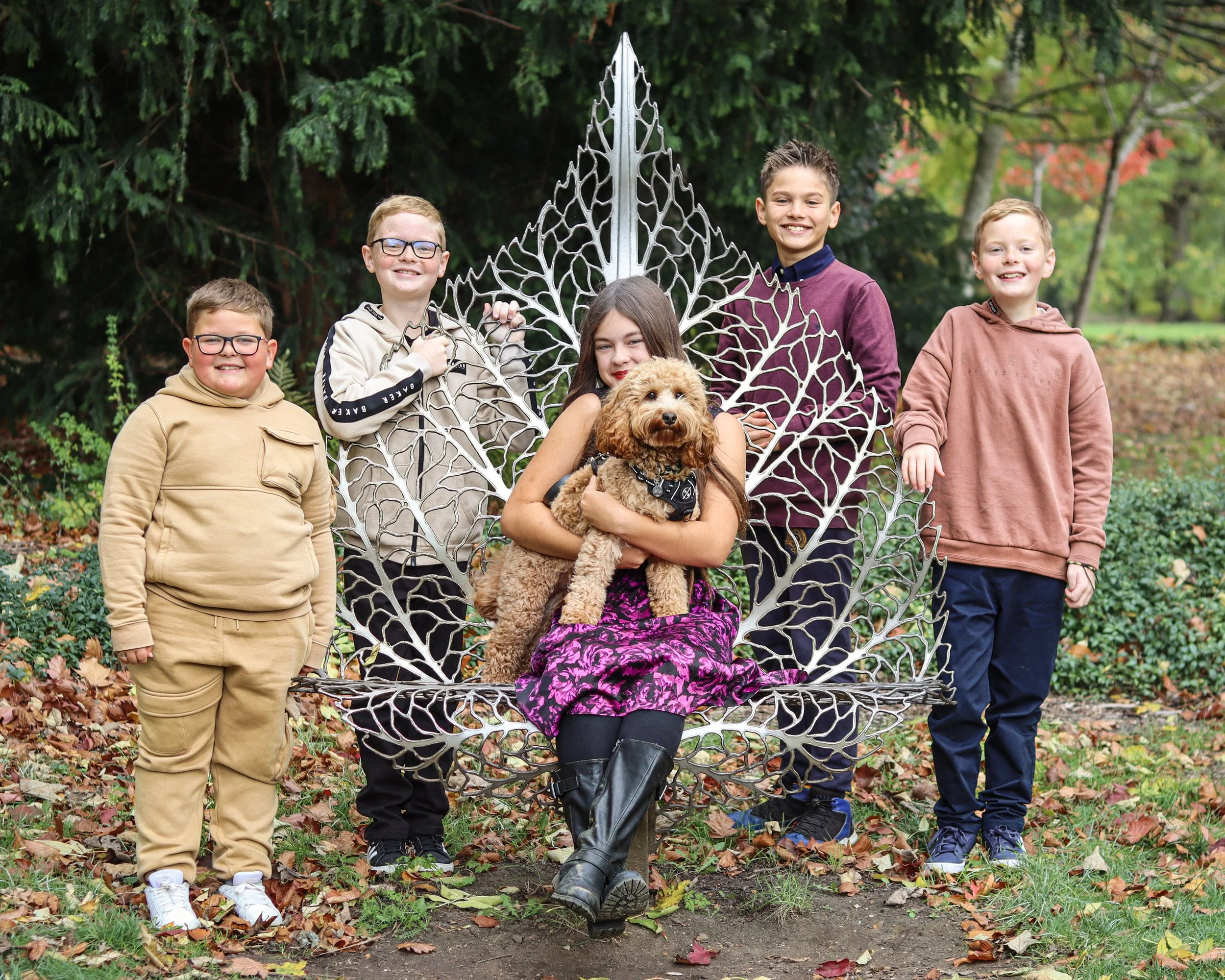 A girl with long brown hair sitting on a decorative metal leaf-shaped bench holding a brown curly-haired dog, with five boys around her smiling outdoors among fall leaves and trees.