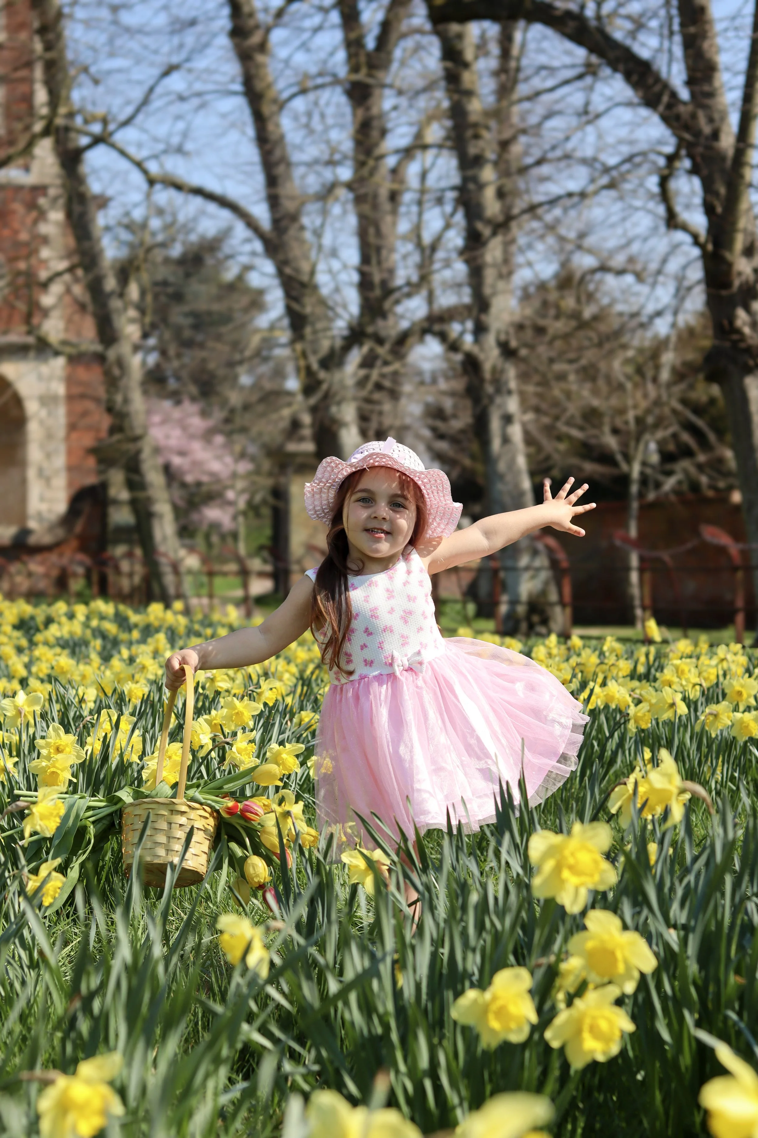 A young girl in a pink dress and sunhat happily playing in a field of yellow flowers, with trees and a building in the background.