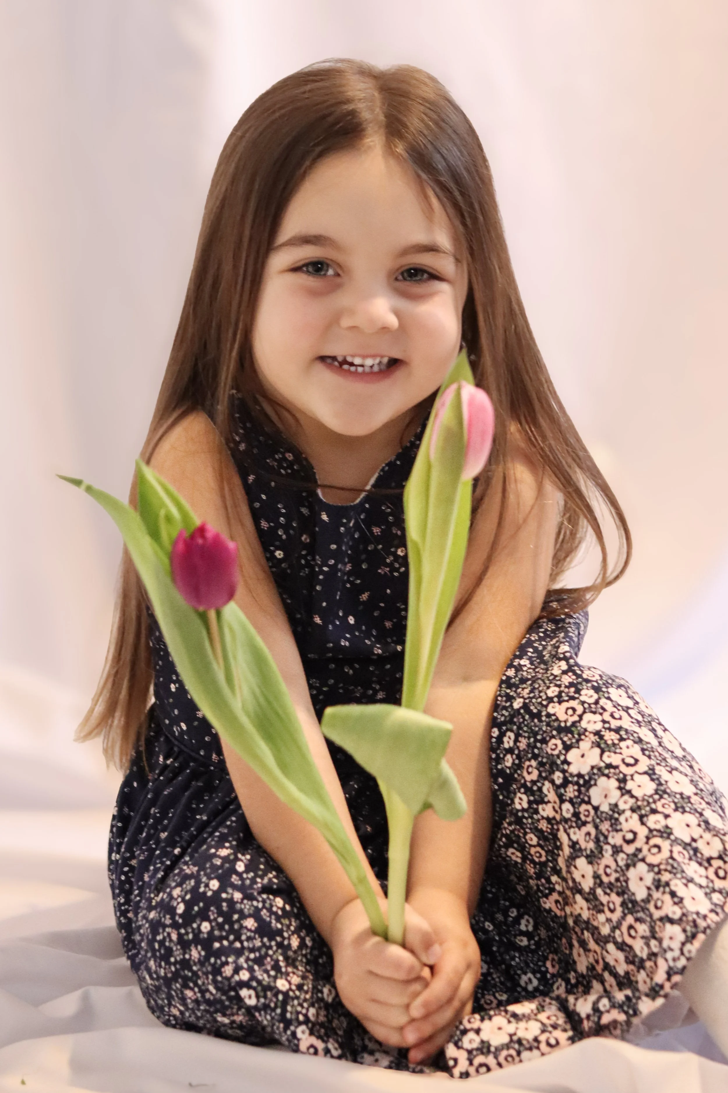 A young girl with long brown hair, wearing a dark floral dress, sitting on a white surface, holding pink and purple tulips, smiling at the camera.