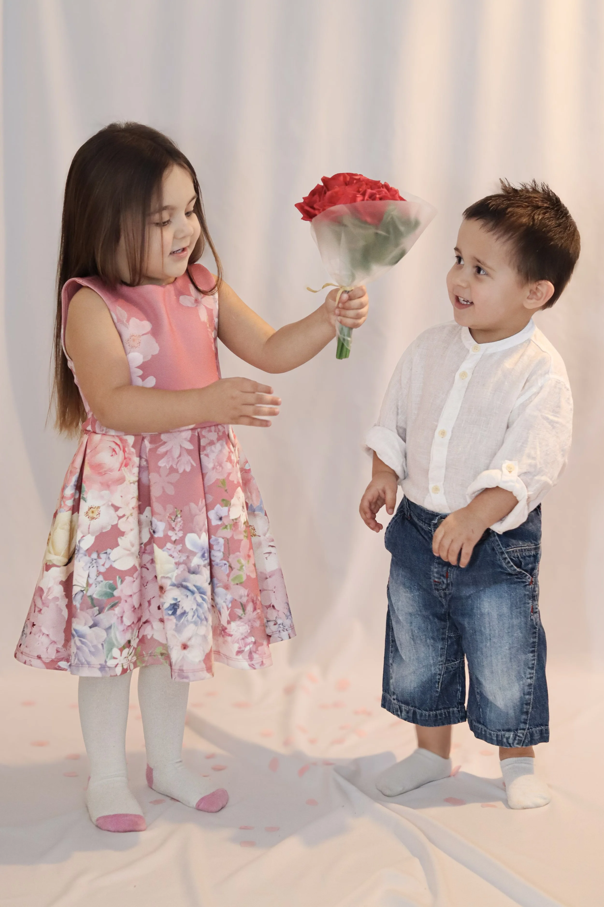 A girl in a floral dress hands a bouquet of red roses to a boy with rolled-up sleeves and denim shorts, on a white background.