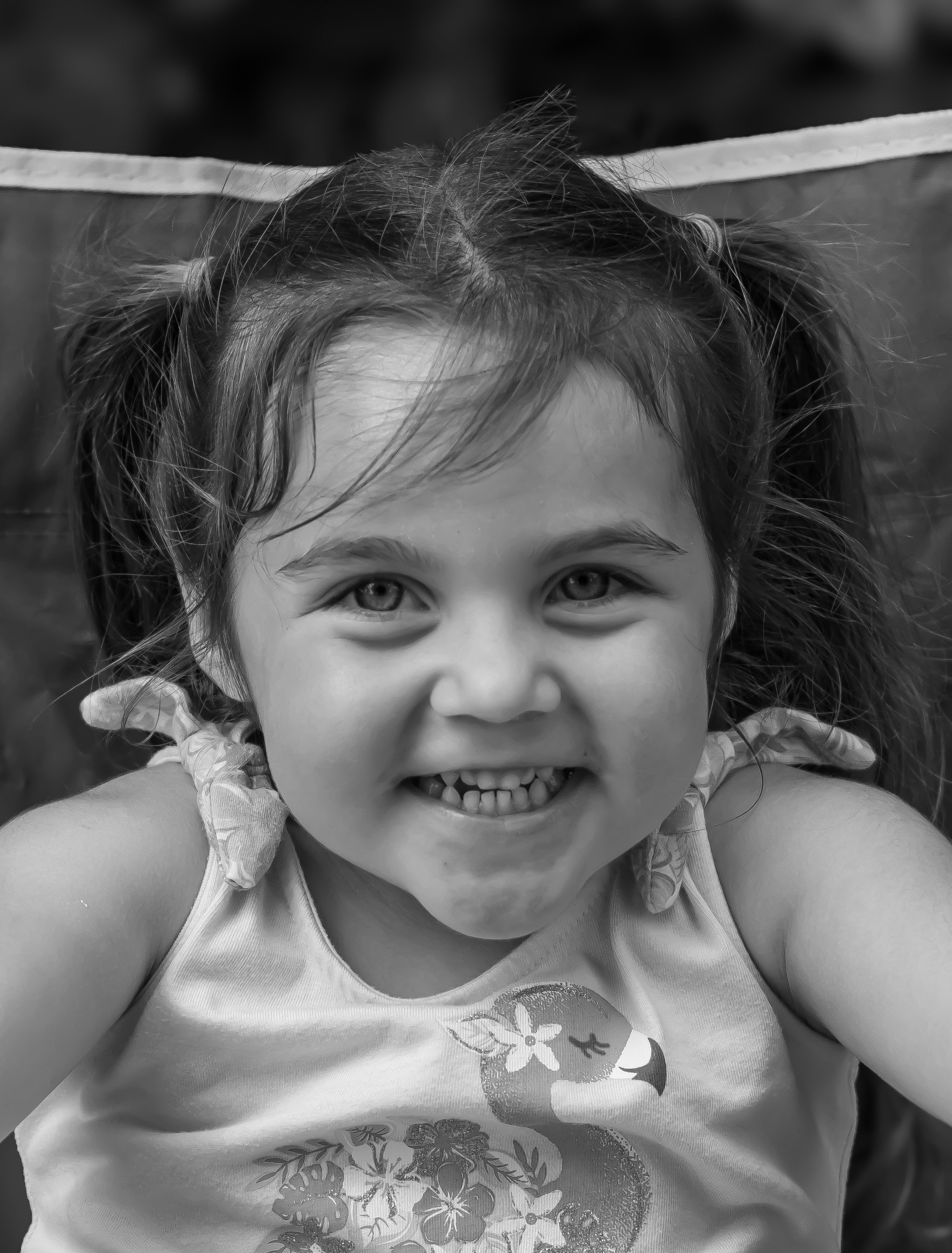 Close-up of a young girl smiling, with her teeth showing, wearing a tank top with a bird and floral design, and tied ribbons in her hair.
