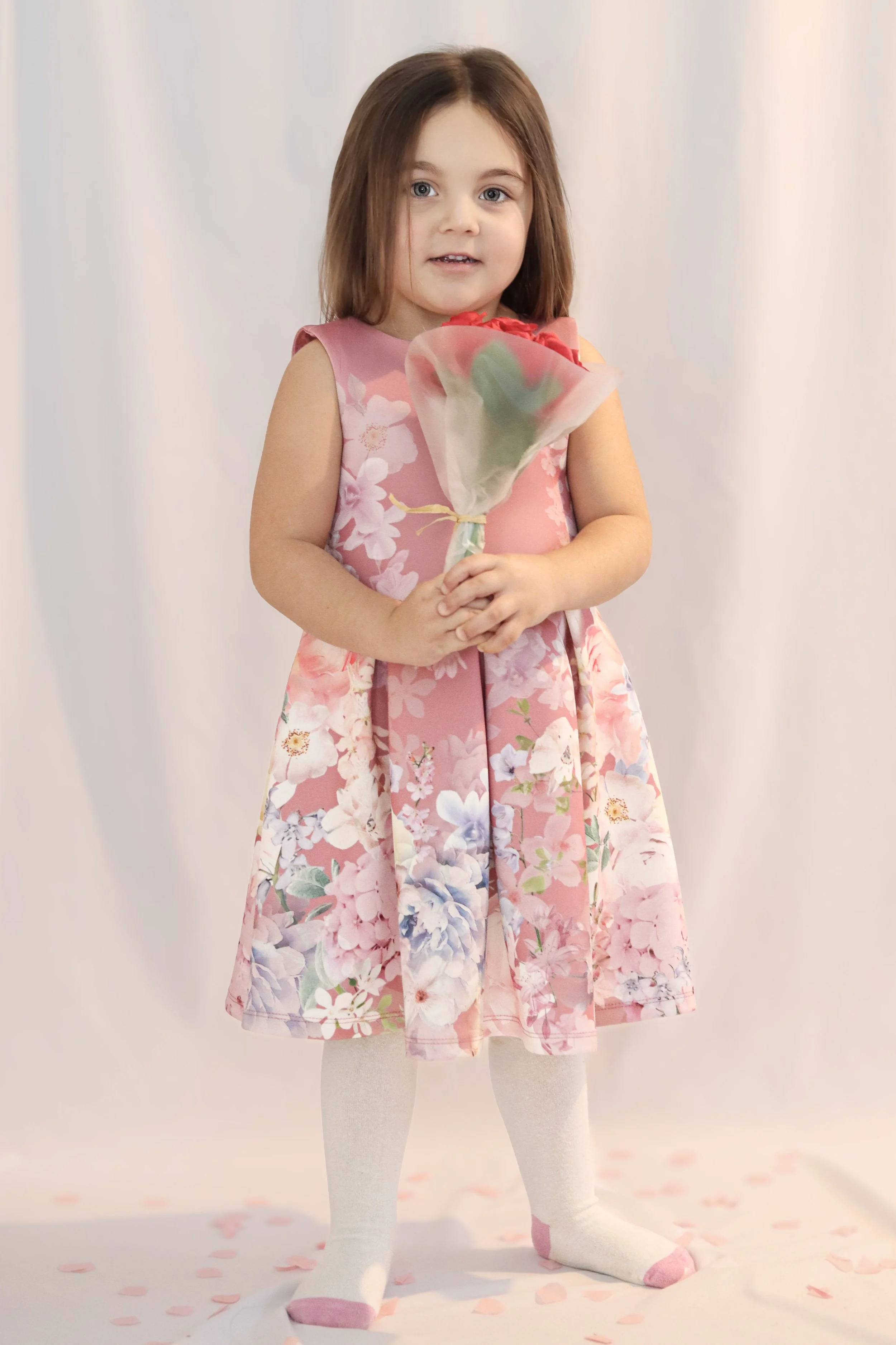 Young girl with shoulder-length brown hair holding a bouquet of flowers, wearing a pink floral dress, white tights, and pink and white socks, standing on a white background with pink flower petals.