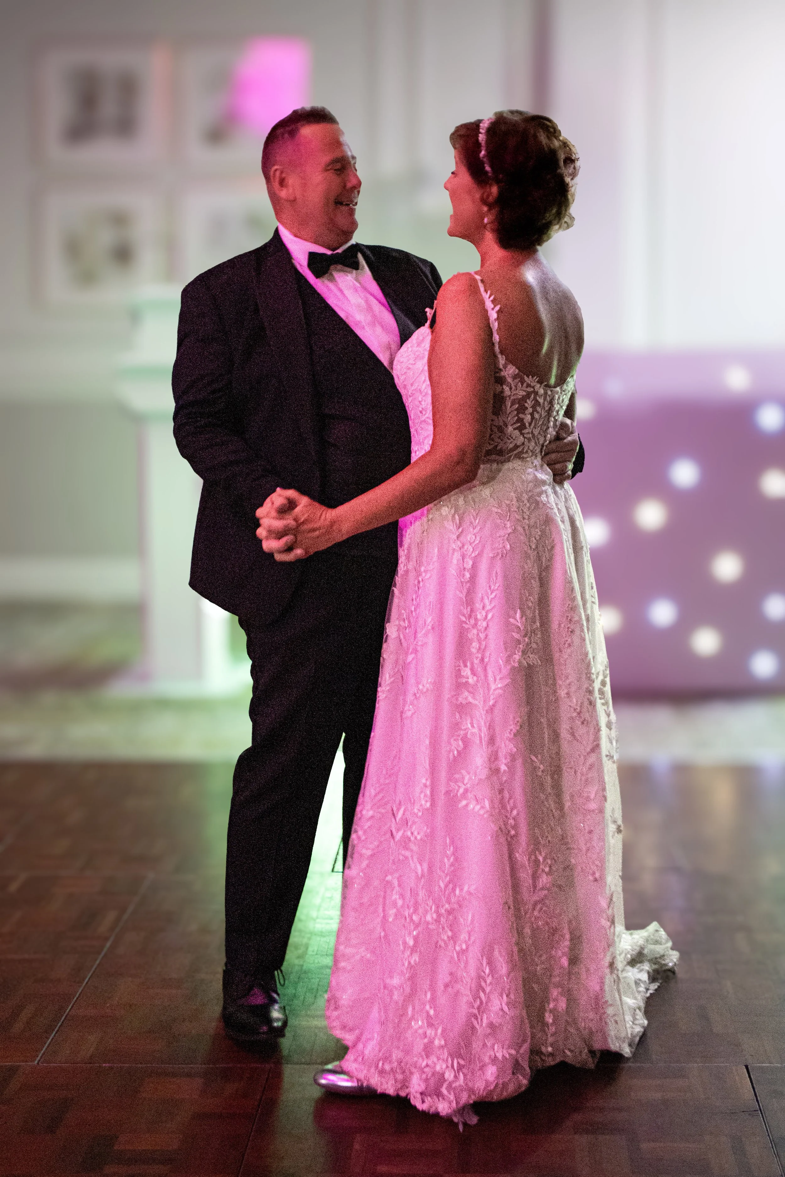 A couple dancing at a wedding reception, the man in a black tuxedo and the woman in a white lace wedding gown.