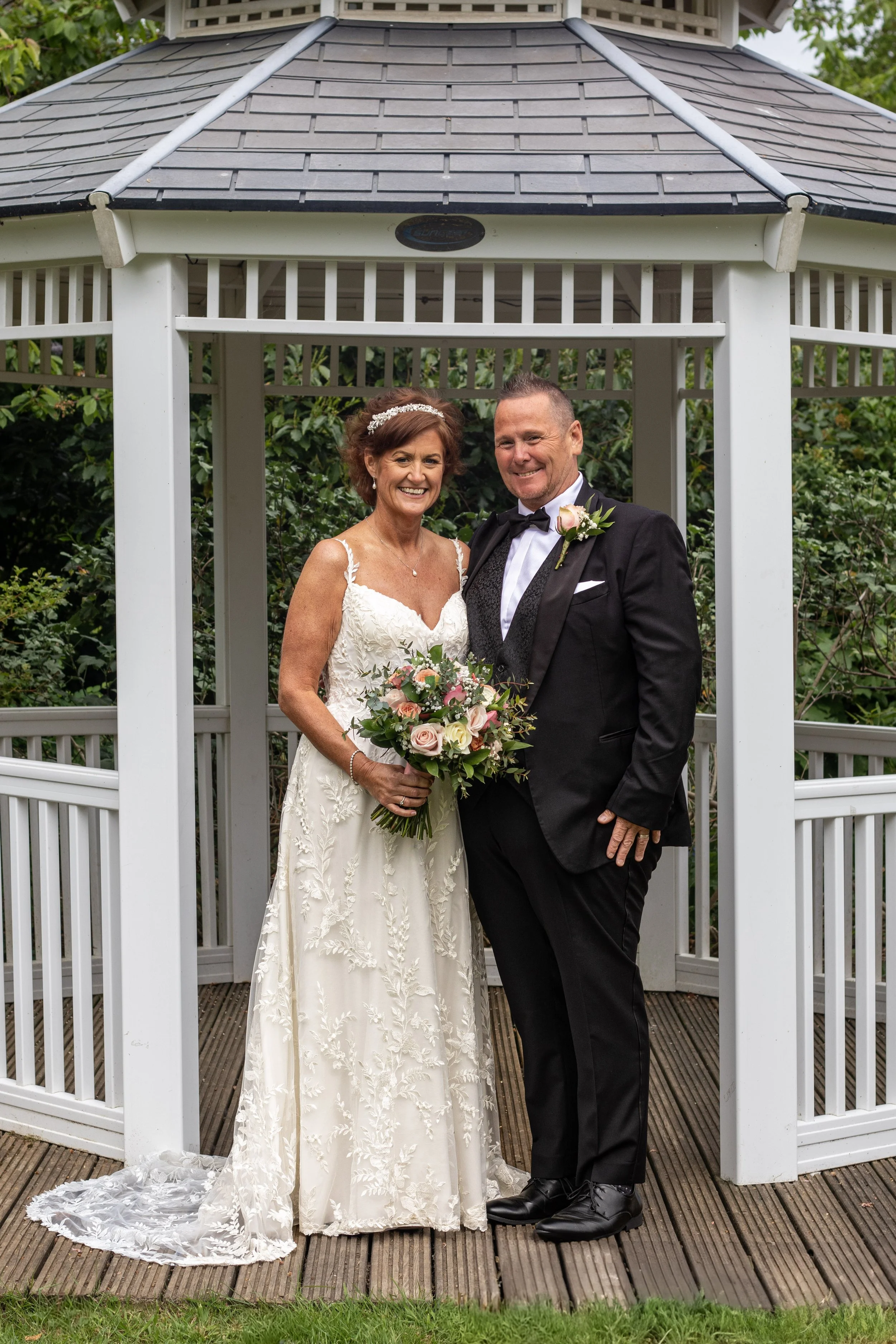 A bride and groom standing inside a white gazebo, smiling and posing for a wedding photo. The bride is holding a bouquet of pink and white flowers, wearing a white lace wedding dress. The groom is dressed in a black tuxedo with a bow tie and a boutonniere.