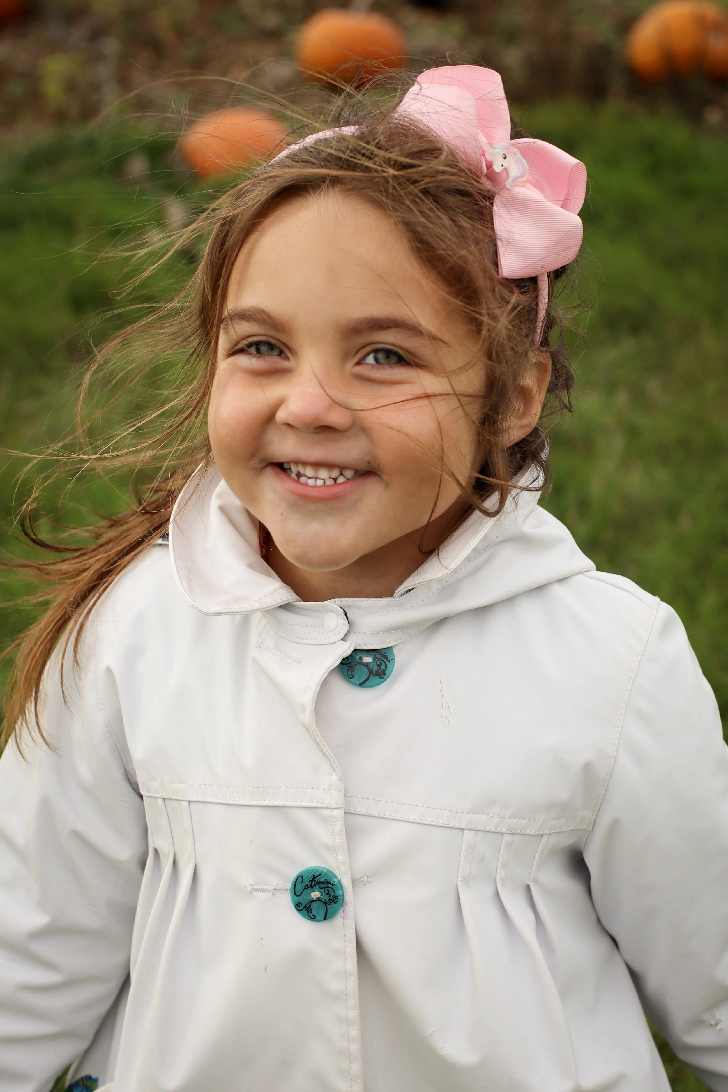 A young girl smiling outdoors with pumpkins in the background, wearing a pink bow in her hair and a white jacket.