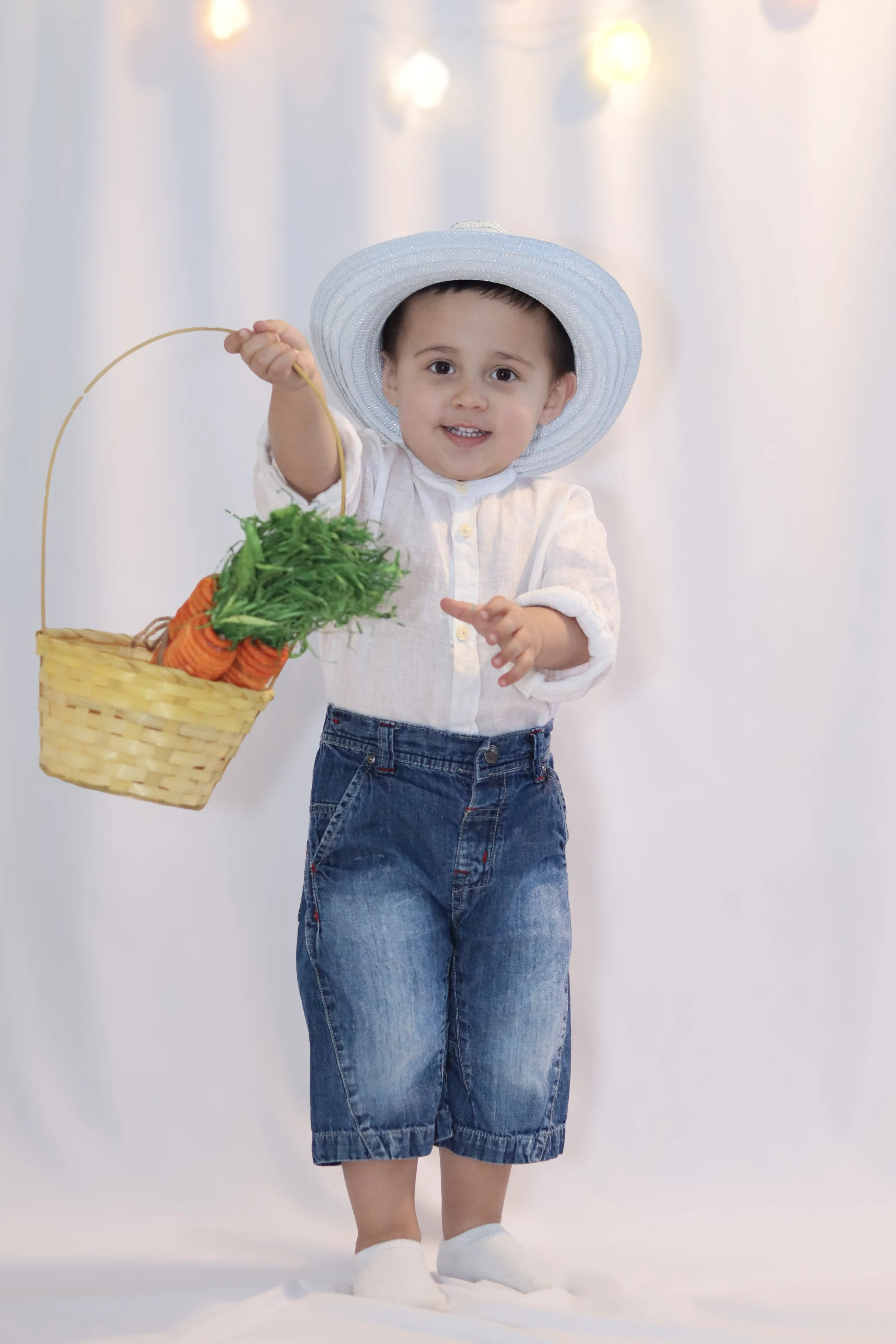 A young child dressed in a white shirt, denim shorts, and white shoes, wearing a large white sun hat, standing against a blurred background with string lights. The child is holding a basket of vegetables, including carrots and leafy greens, and smiling at the camera.