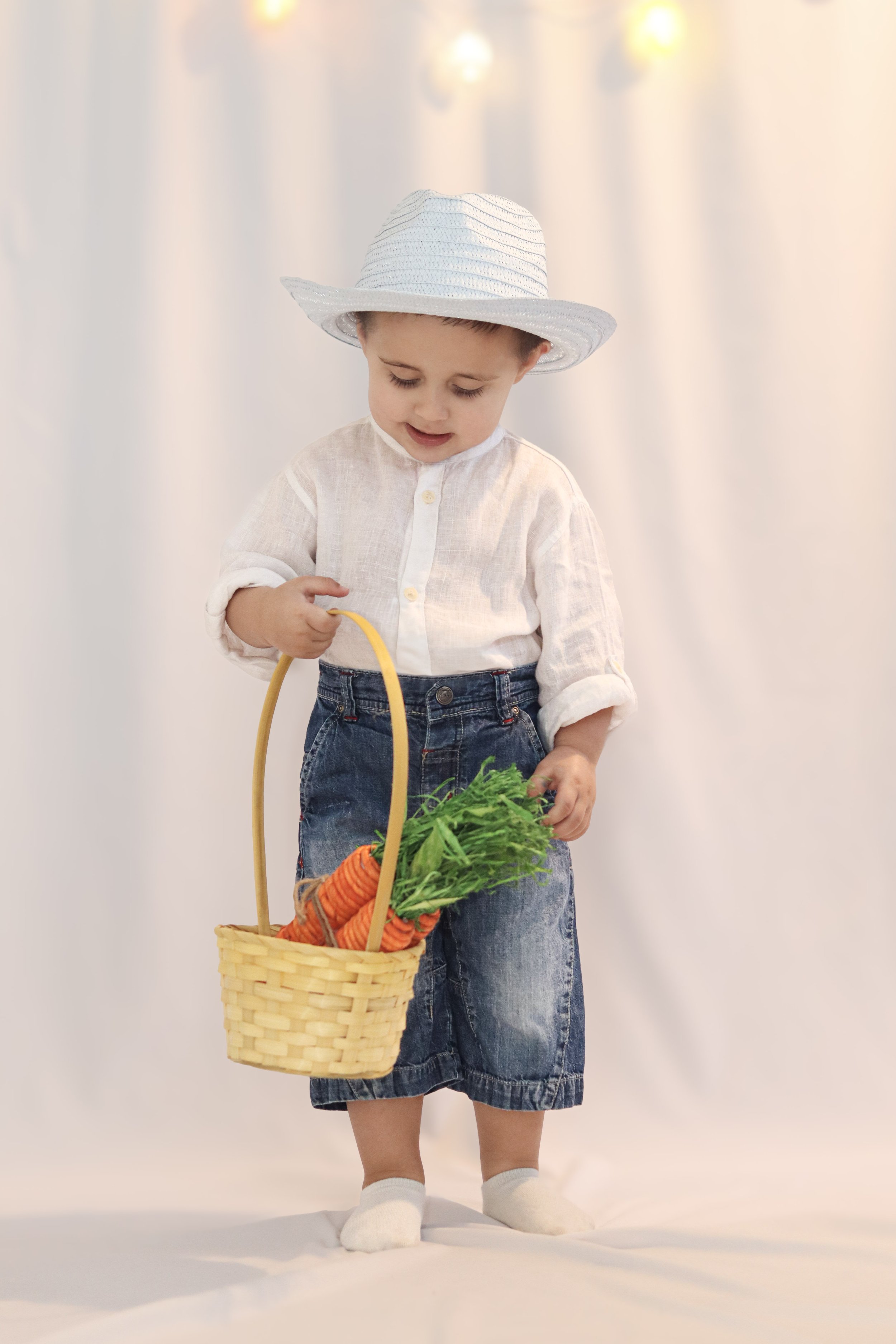 A young child wearing a white hat, white shirt, and denim shorts holding a basket of carrots and looking down at them.