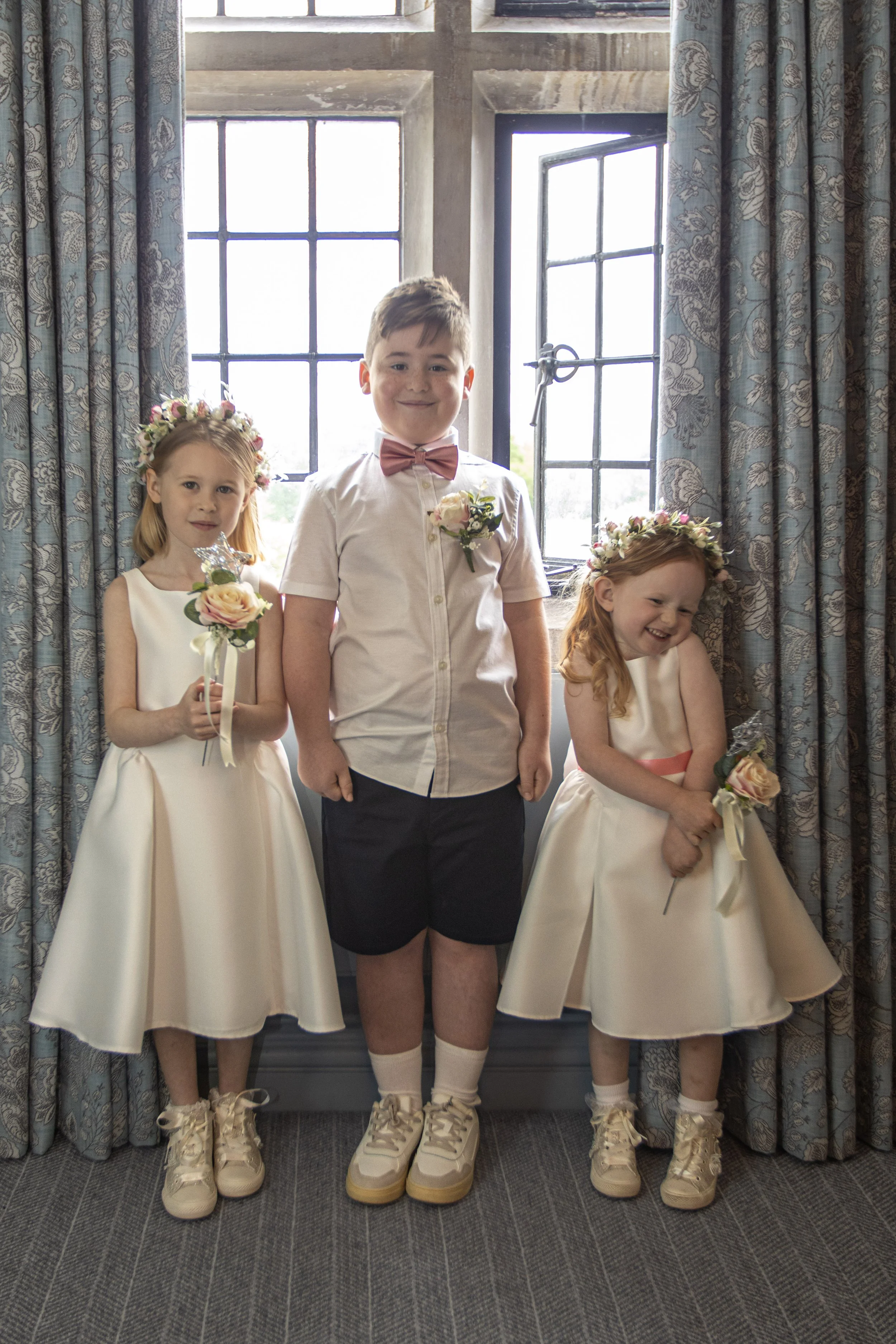 Three children dressed in formal attire standing in front of an open window with blue patterned curtains; the two girls are wearing cream-colored dresses and floral crowns, holding bouquets of flowers, and the boy is wearing a white shirt, pink bow tie, and dark shorts.