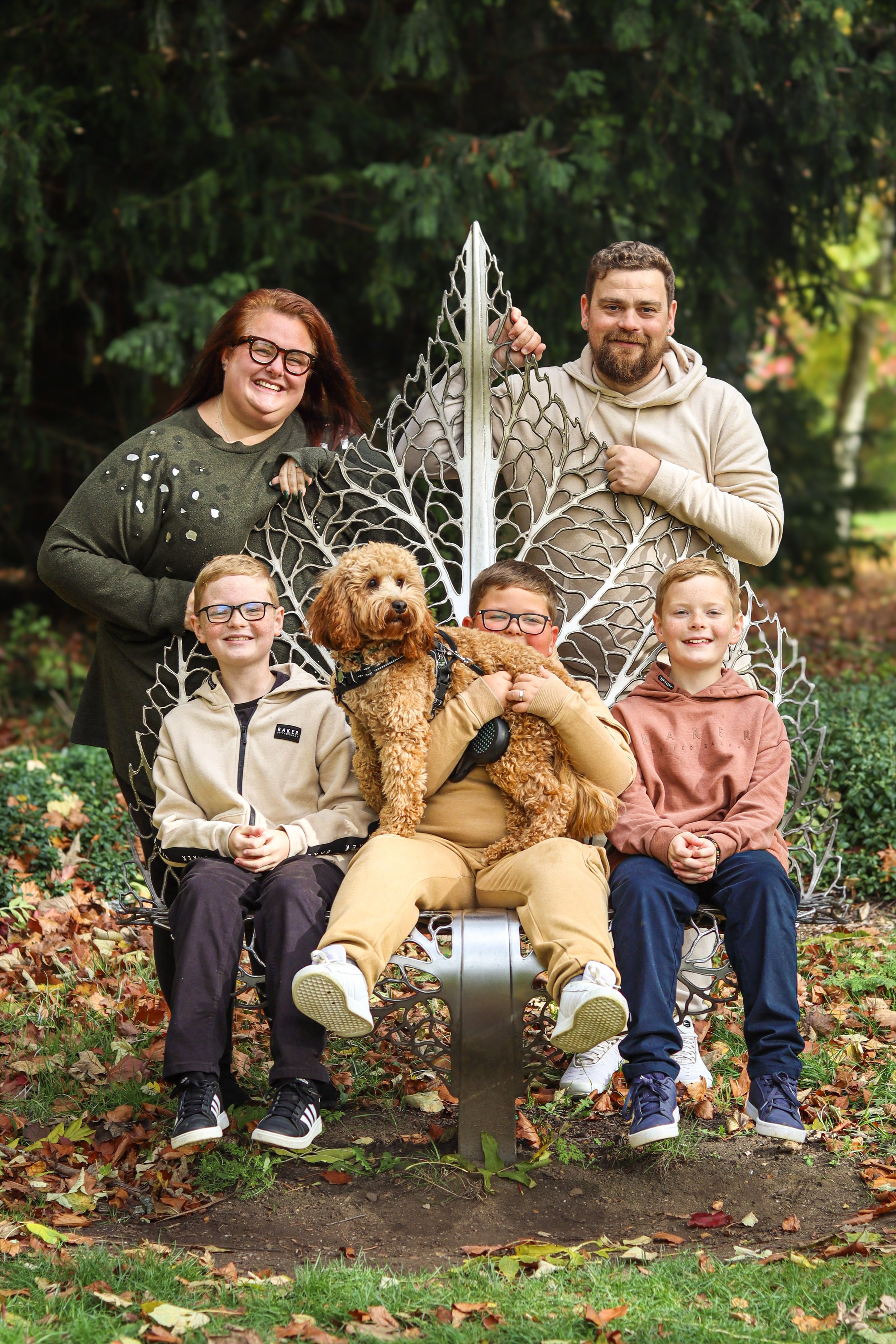 A family of six, including four children, two adults, and a dog, sitting and standing around a decorative metal bench with leaf designs in a park during autumn. The children are seated and the adults are standing, all smiling at the camera.