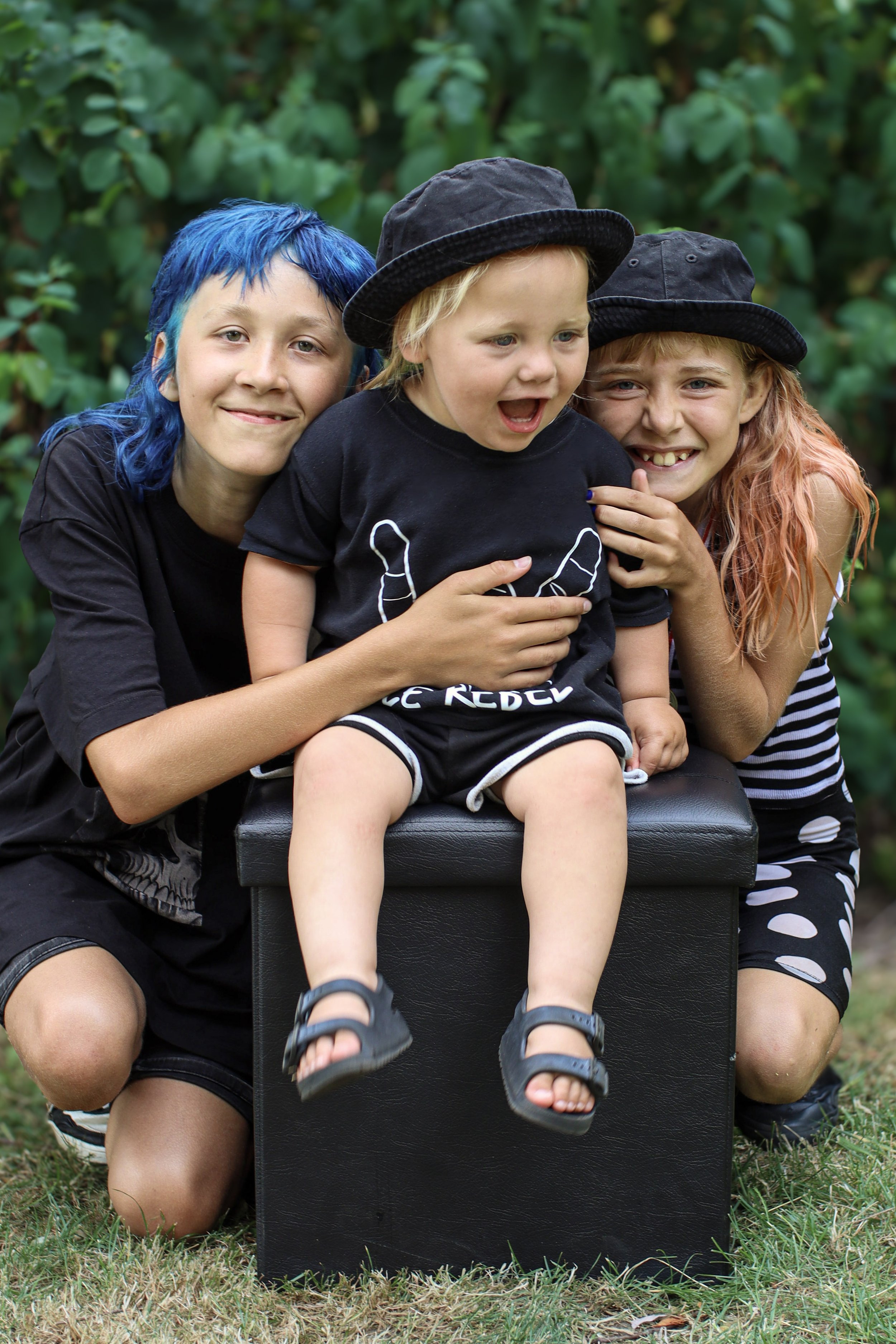 Three children with colorful hair posing outdoors, one sitting on a black box and two kneeling beside it, all smiling and wearing black hats and casual clothes, surrounded by greenery.