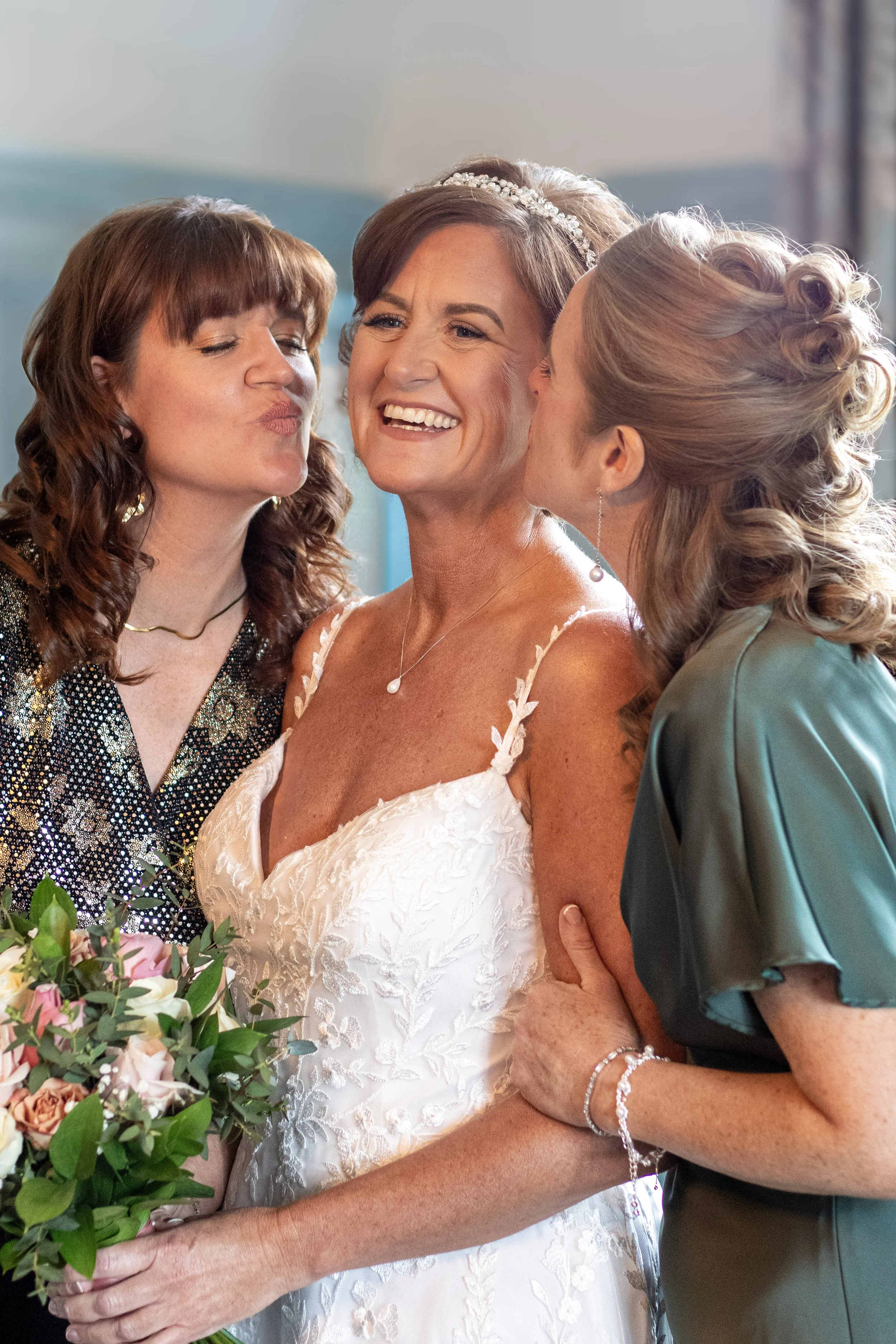A bride in a white wedding dress with a veil, holding a bouquet of flowers, is surrounded by two women who are kissing her on the cheeks during her wedding celebration.