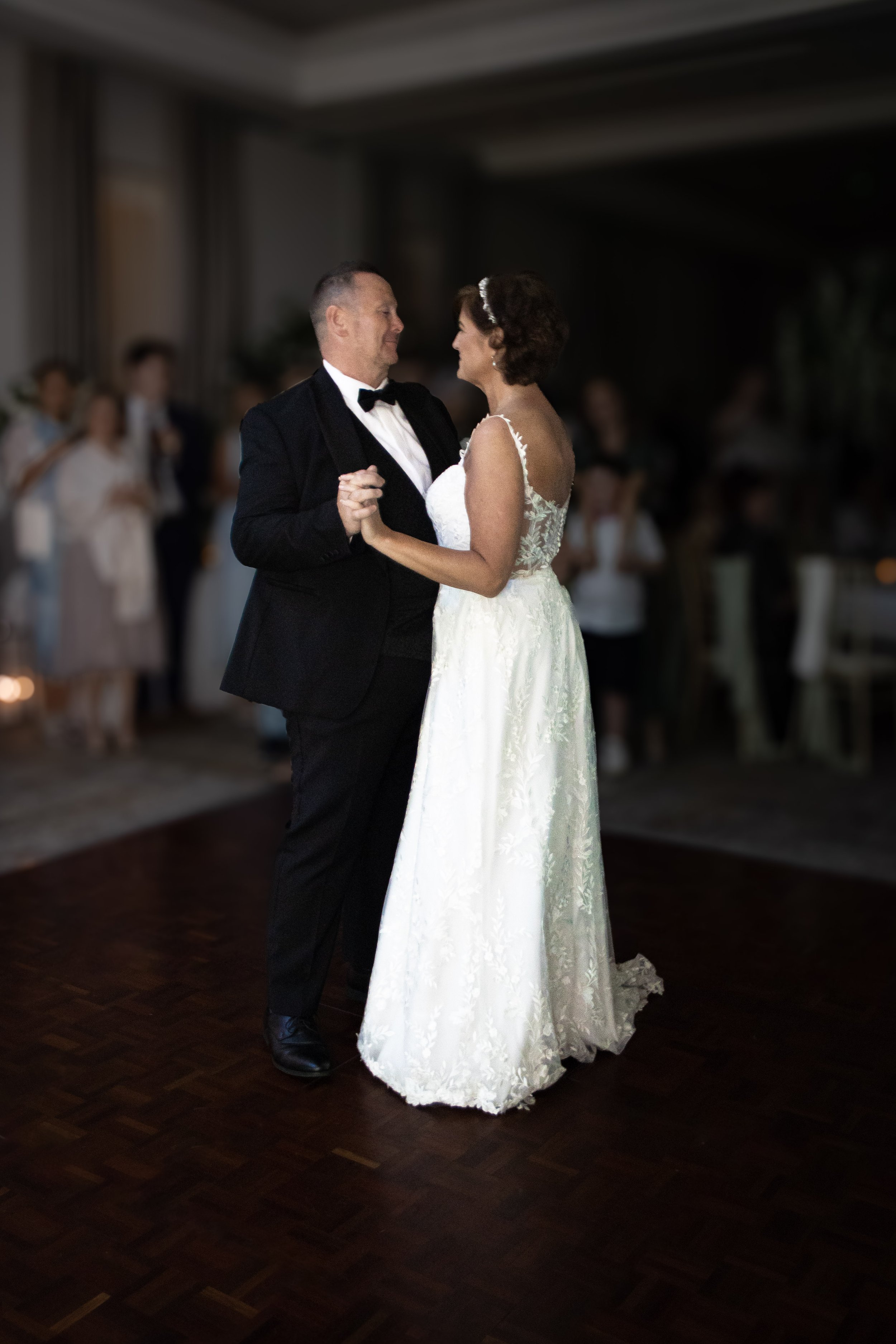 A bride and groom are dancing at their wedding reception, surrounded by guests, in a dimly lit indoor venue.