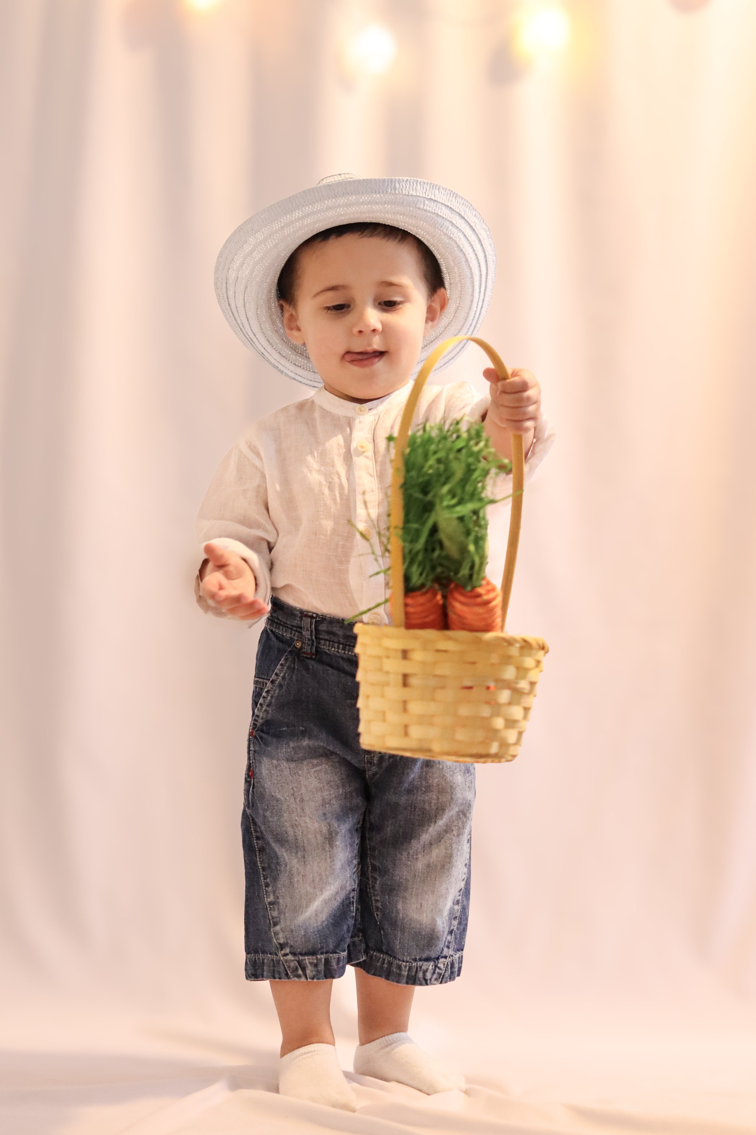 A young boy wearing a large white sun hat, beige shirt, denim shorts, and white socks, holding a wicker basket filled with vegetables including carrots and leafy greens.