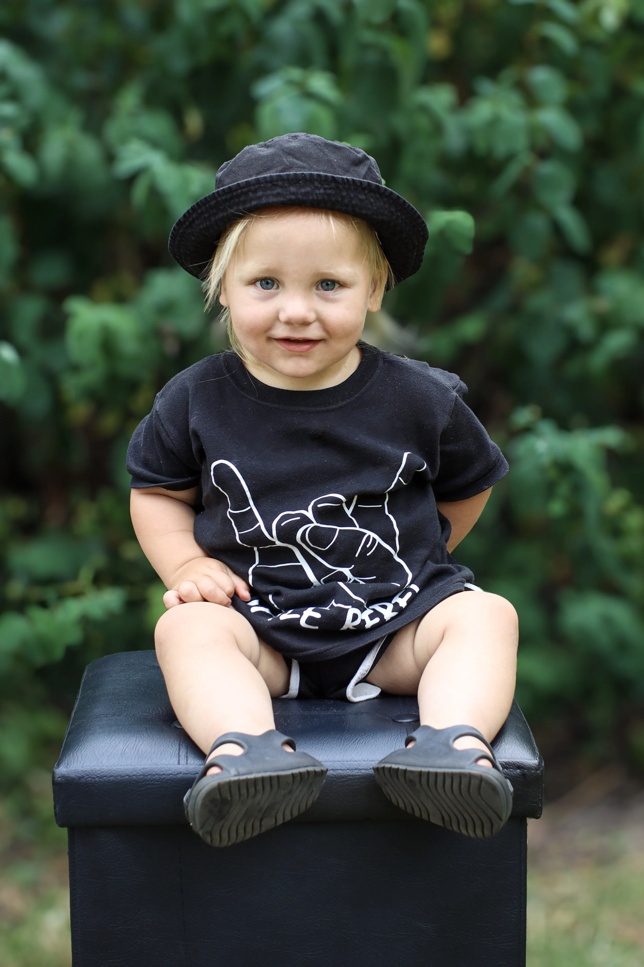A young boy with blonde hair and blue eyes sitting on a black box outdoors, wearing a black t-shirt with a white graphic, a black hat, and black sandals with socks, surrounded by green foliage.