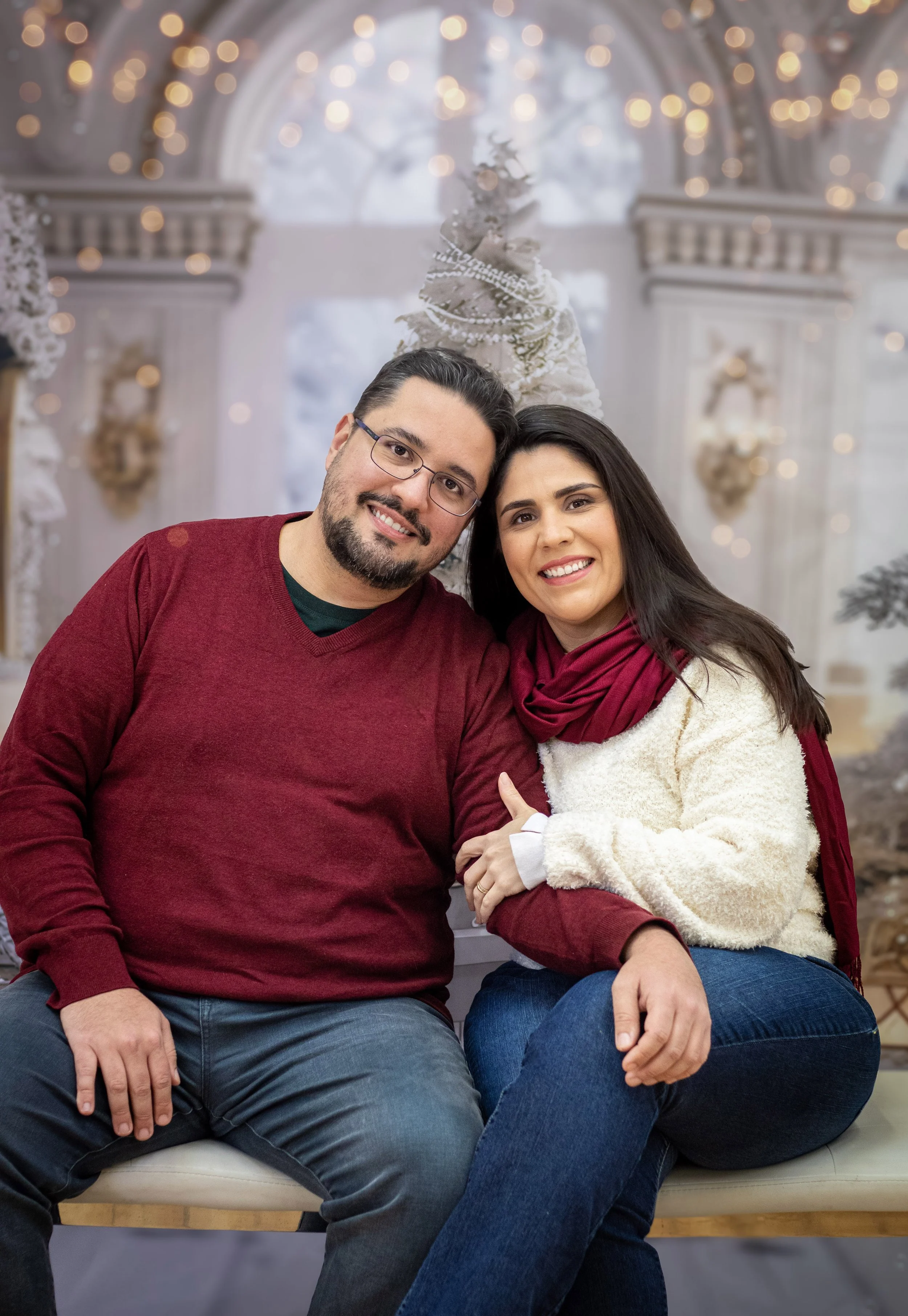 A smiling couple sitting close together on a bench, embracing each other in a festive, Christmas-themed setting with a decorated Christmas tree and twinkling lights in the background.