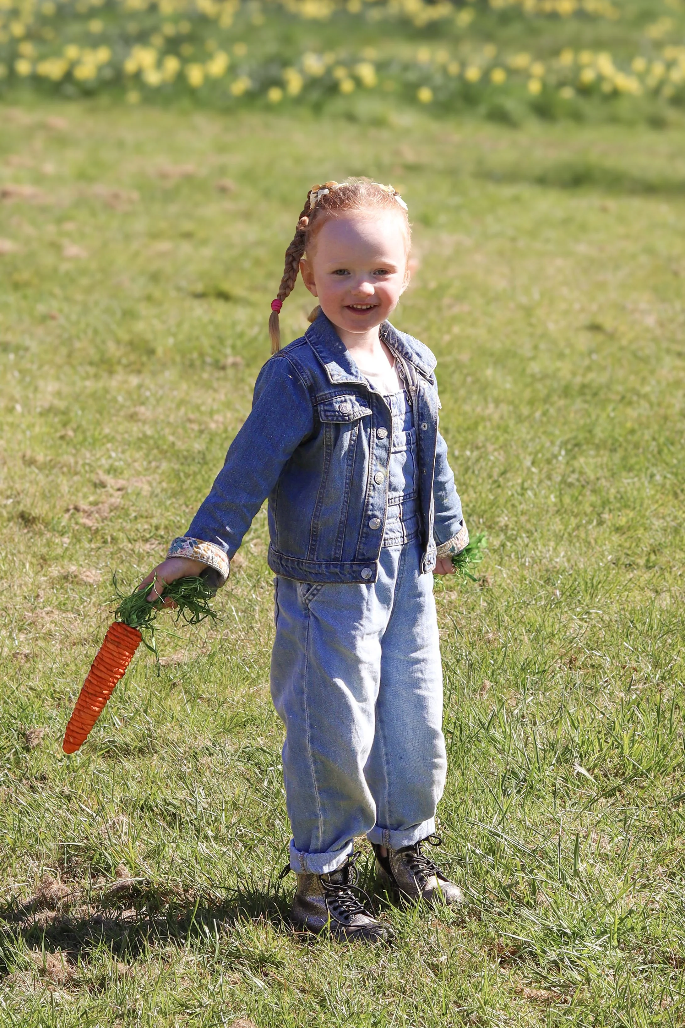 A young girl with red hair in braids, wearing a denim jacket and jeans, holding a large artificial orange carrot in a grassy field with yellow flowers in the background.