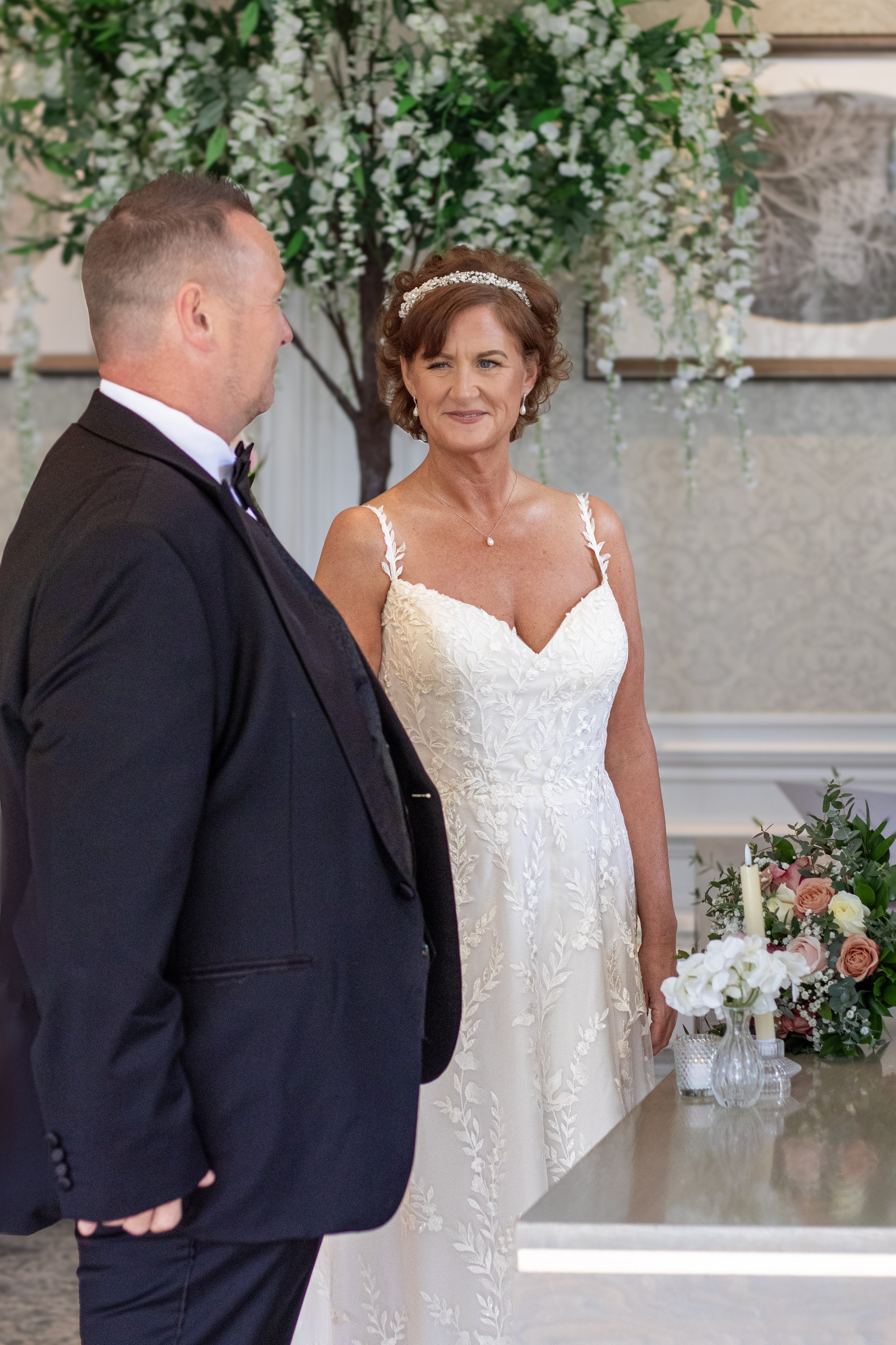 A bride and groom stand facing each other during a wedding ceremony indoors, with floral arrangements and greenery in the background.