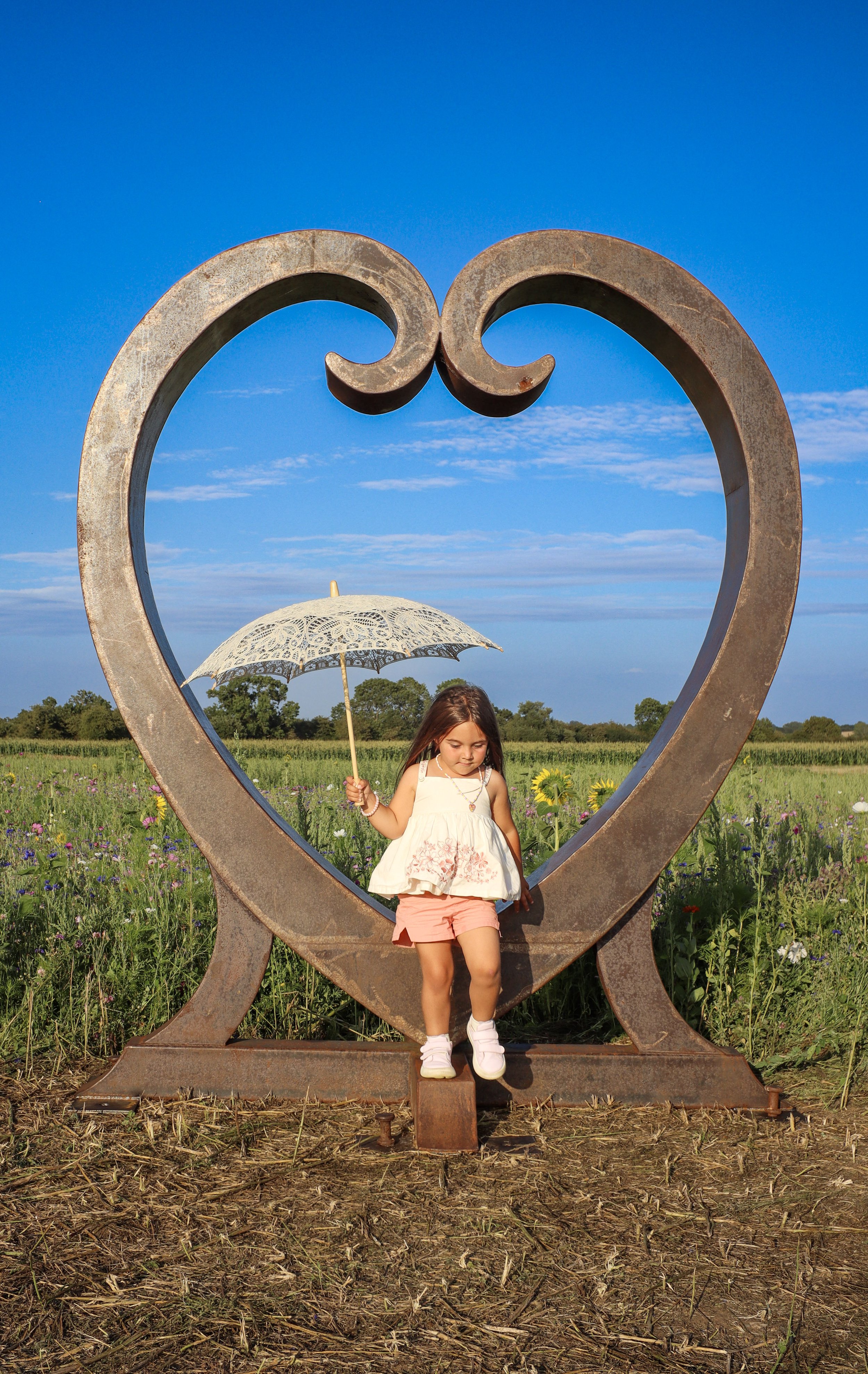 A young girl standing inside a large, rusted heart-shaped metal sculpture in a field of wildflowers under a blue sky, holding a white lace umbrella.