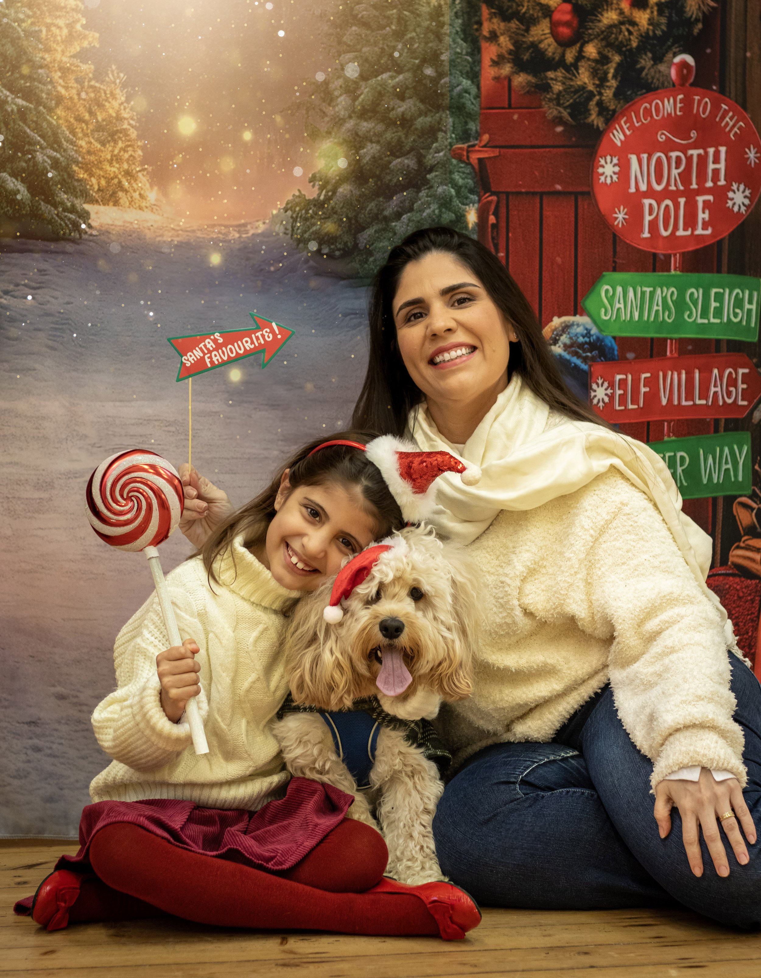 A woman, a girl, and a dog wearing Santa hats posing indoors with Christmas decorations and signs in the background, holiday-themed setting with snow, trees, and festive signage.