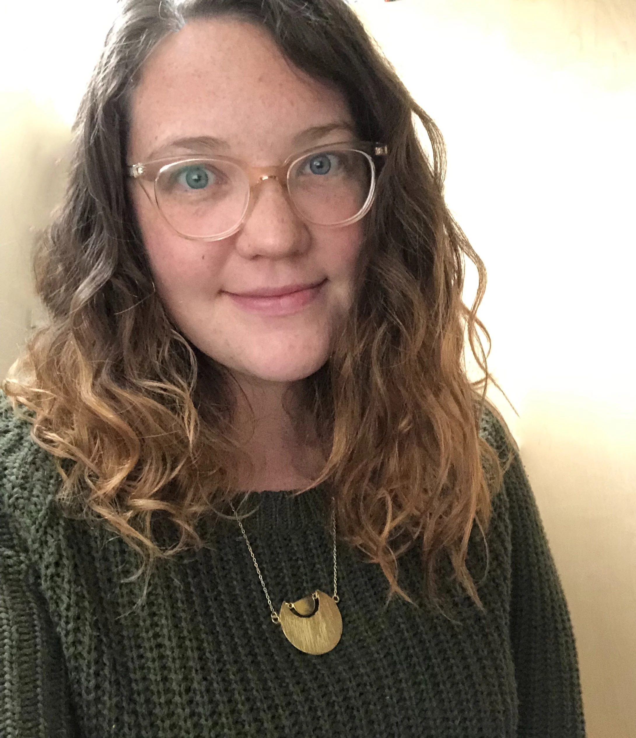 Person with wavy hair wearing glasses, a dark green sweater, and a gold necklace, smiling at the camera.