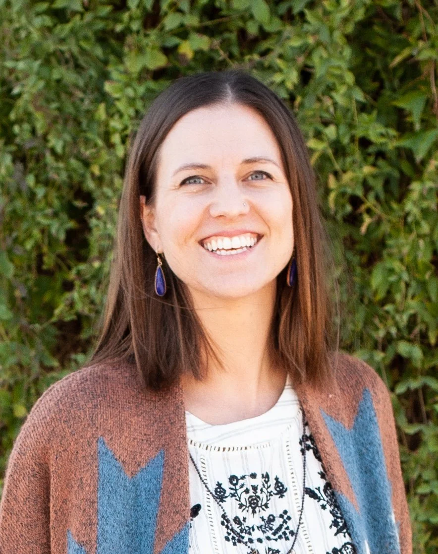 A woman with shoulder-length brown hair smiling outdoors in front of green leafy bushes, wearing a brown cardigan with blue accents over a white embroidered blouse, accessorized with dangling earrings and a necklace.