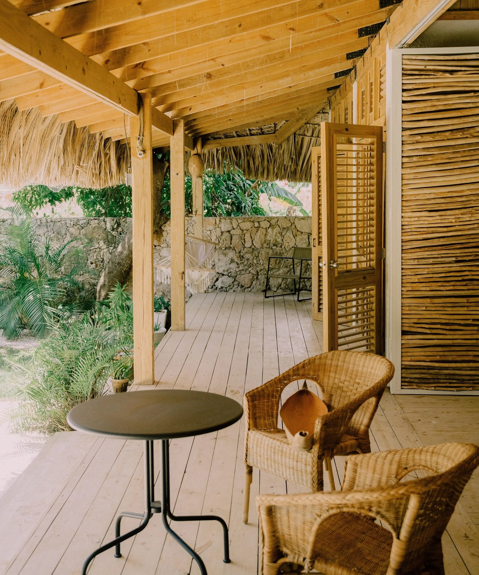 A porch with a wooden floor and ceiling, surrounded by greenery, with wicker chairs, a small black table, a hammock, and a stone wall in the background.