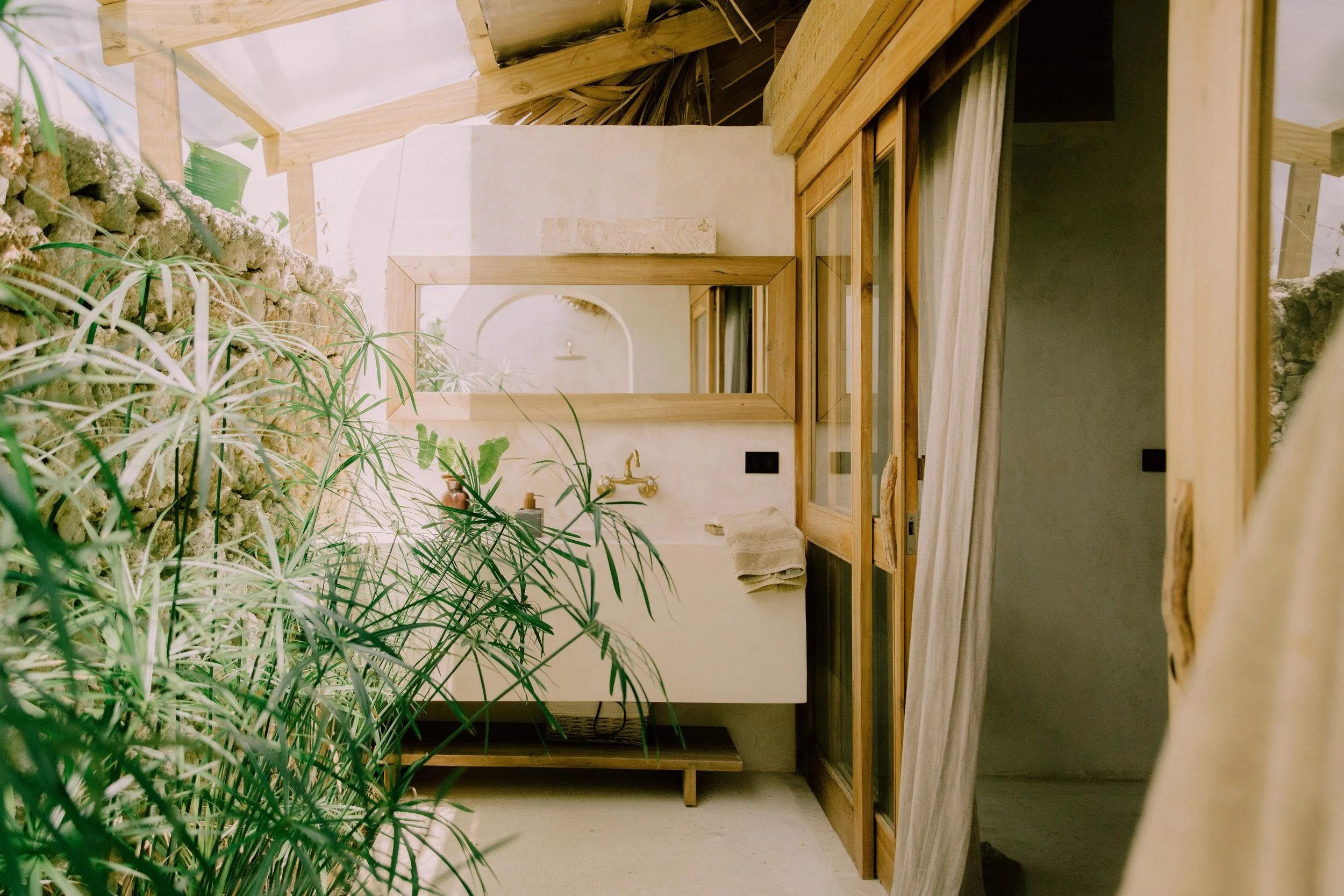 Un espacio interior con paredes de piedra y madera, una ventana grande con cortinas claras, plantas verdes y un lavabo elegante con accesorios en tonos dorados.