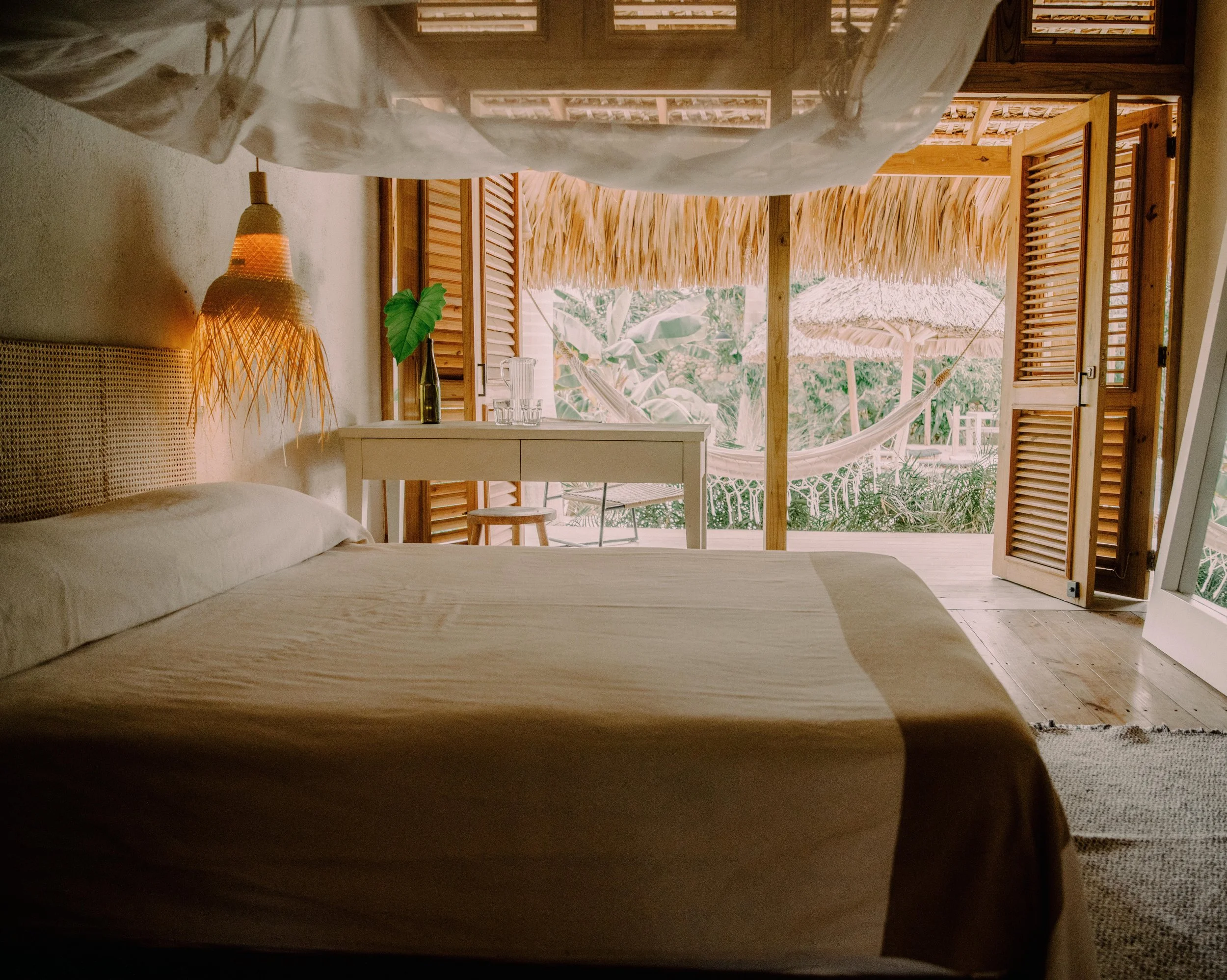 Habitación con cama y vista a un espacio exterior con hamacas, palapas y vegetación tropical.