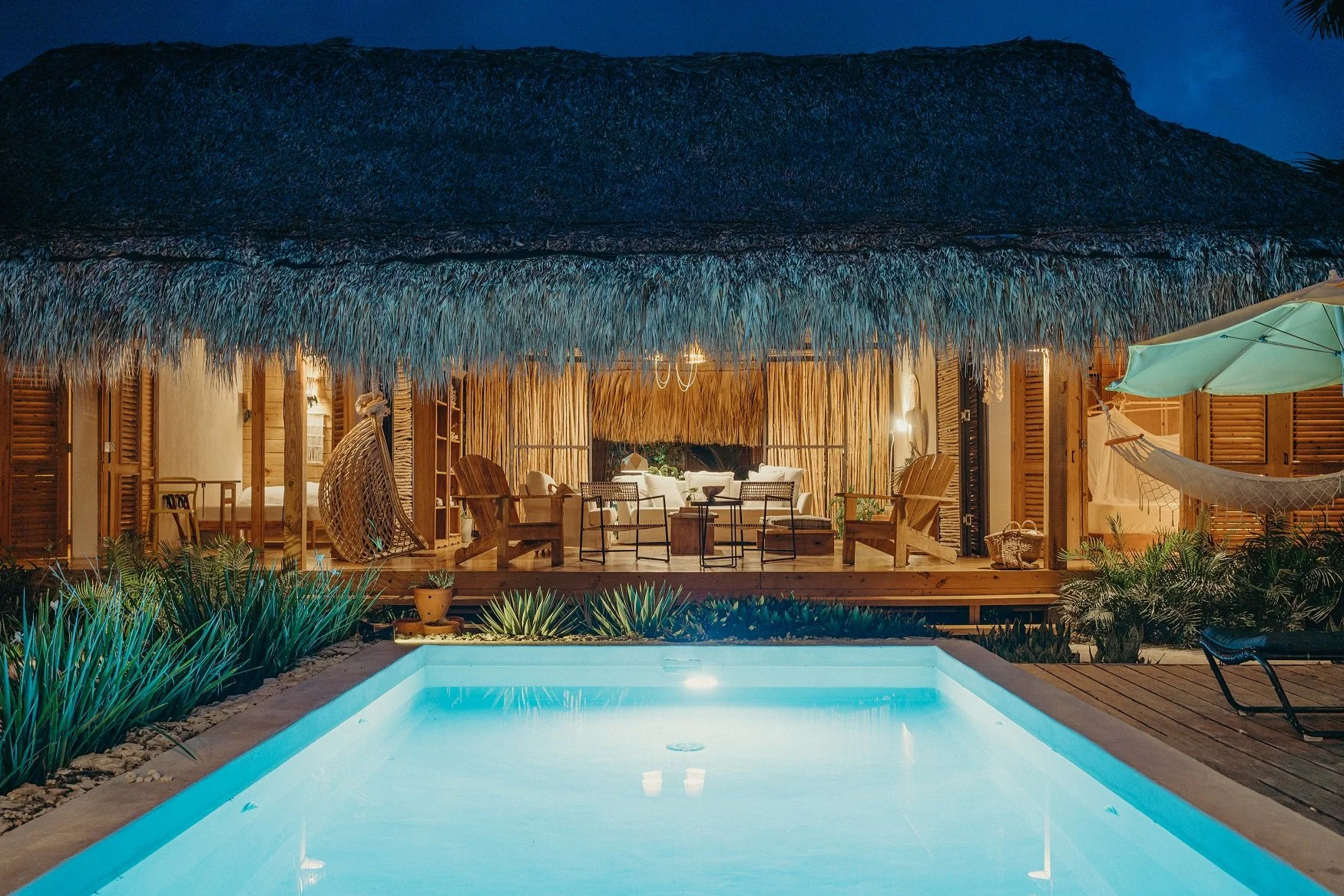 Casa rústica con techo de paja, iluminada desde adentro, con terraza de madera y mobiliario de sala, vista a una piscina iluminada en la noche, rodeada de vegetación tropical y una sombrilla de tela beige a la derecha.