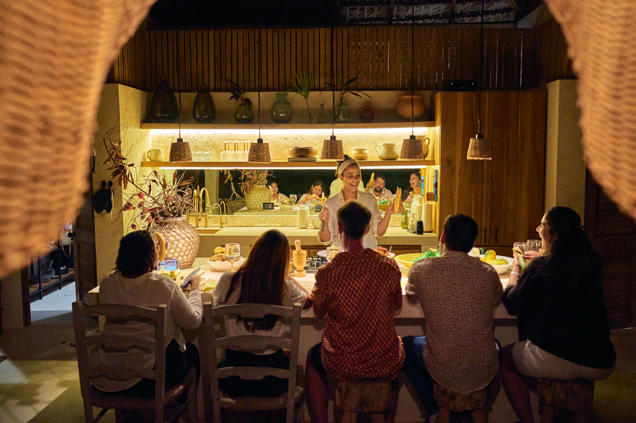 Grupo de personas disfrutando de una comida en una cocina moderna, con una mujer de pie sirviendo comida a los comensales, en un ambiente cálido y bien iluminado.