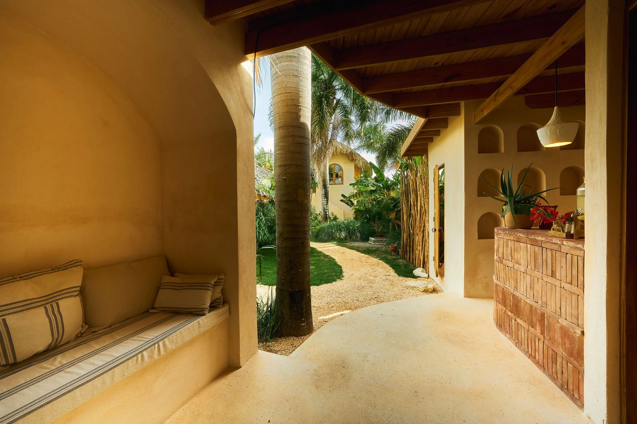 Vista del porche de una casa con paredes amarillas, un árbol de palma y un camino de grava, con plantas tropicales y muebles de madera.