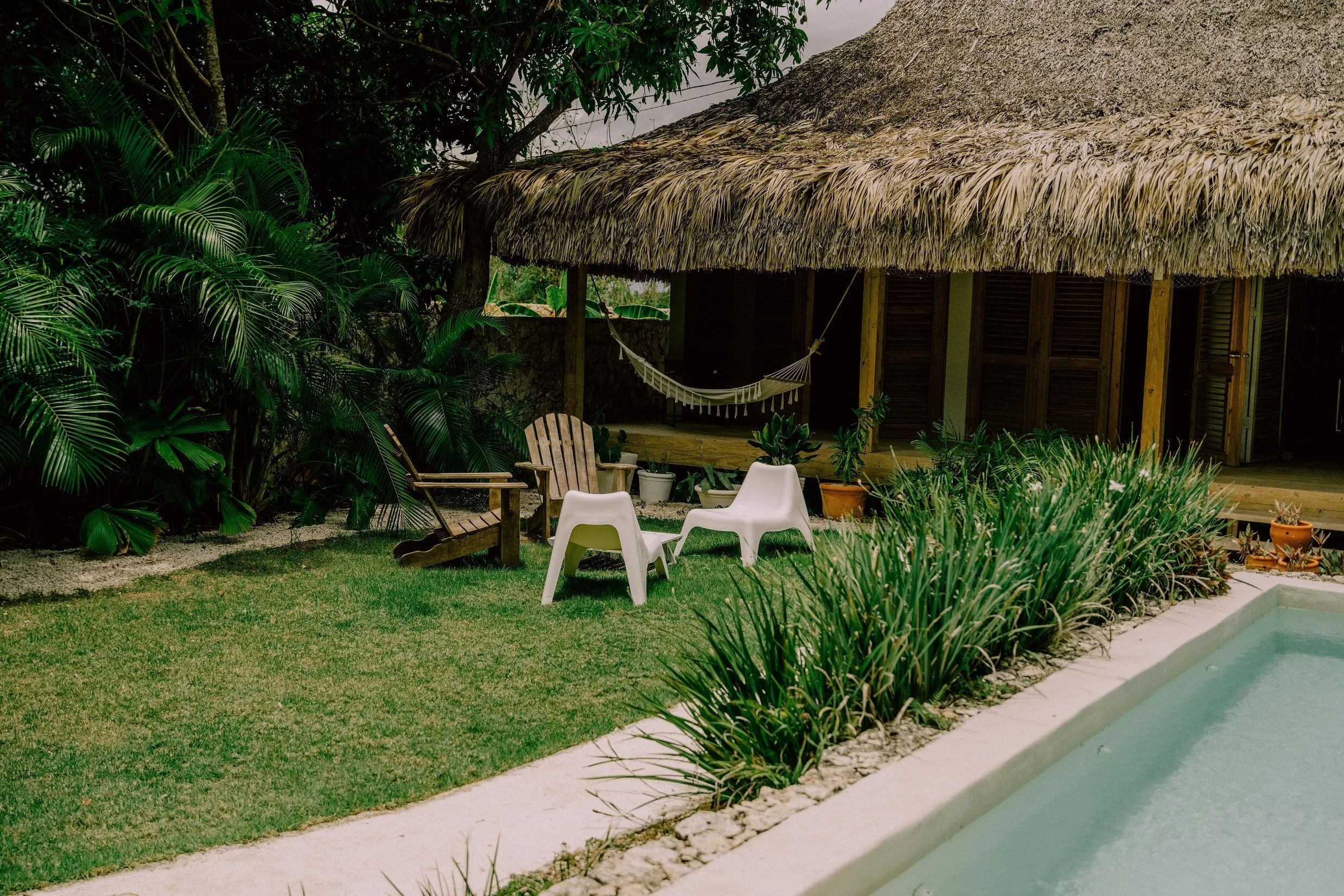 Área exterior con hamaca, sillas de plástico y de madera, plantas y alberca junto a una cabaña con techo de palapa.