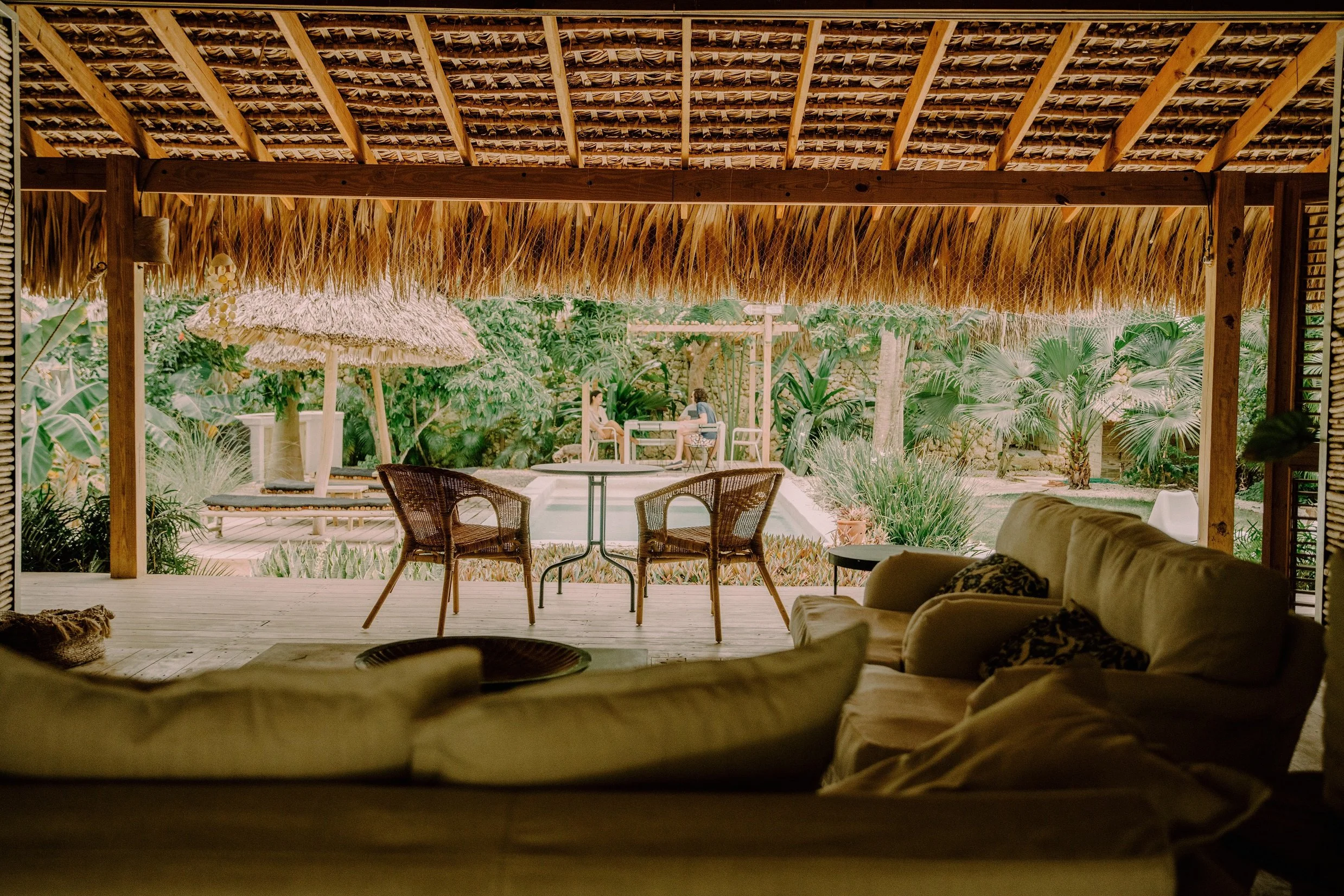 Vista desde una sala con sofá y sillones hacia un área al aire libre con césped, piscina, sillas de mimbre, sombrilla de paja, y vegetación tropical en un entorno de playa o resort.