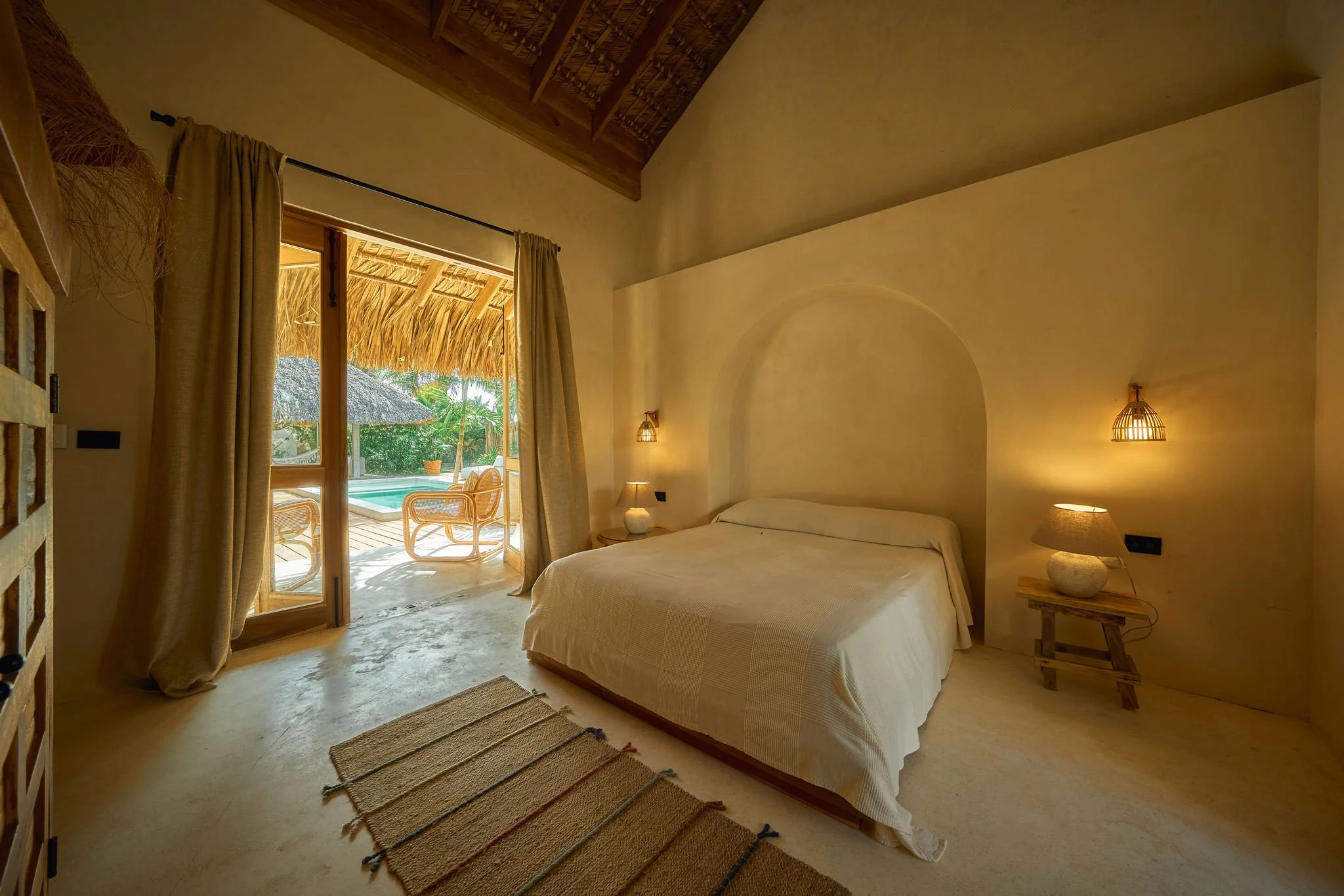 Habitación con cama blanca, lámparas y vista a una terraza con sillas de mimbre y piscina, rodeada de palmeras y vegetación tropical.
