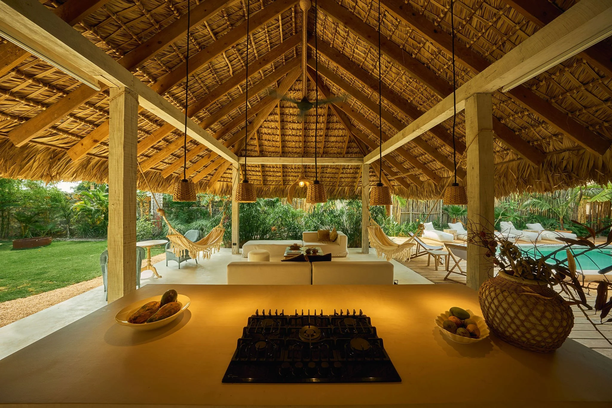 Vista de una cocina y área de descanso en una casa de terraza con techos de paja, muebles blancos y hamacas, rodeada de vegetación tropical, con piscina al fondo.
