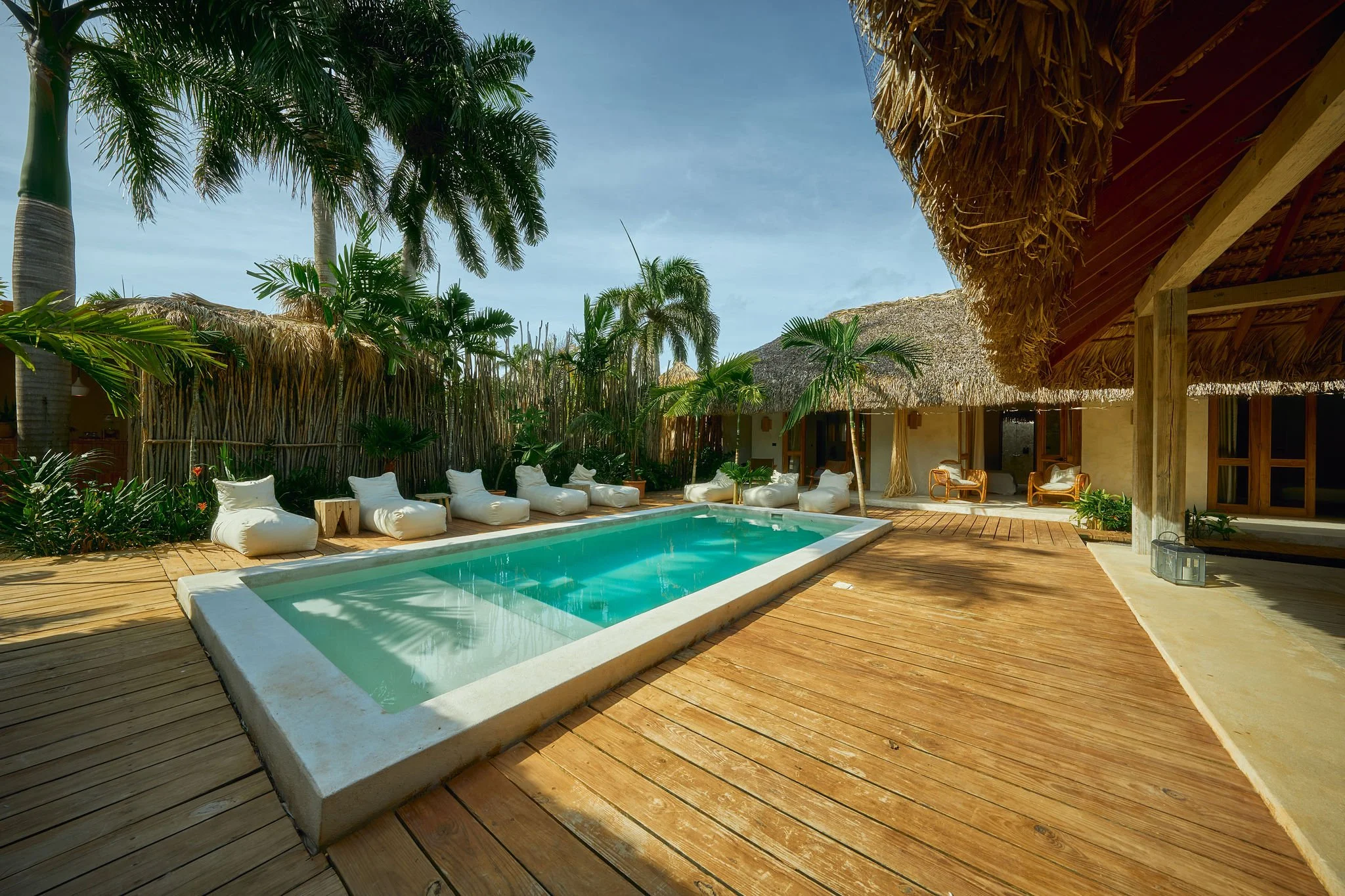 Área de alberca en un resort tropical con tumbonas blancas, palmeras y cabañas de madera con techos de paja.