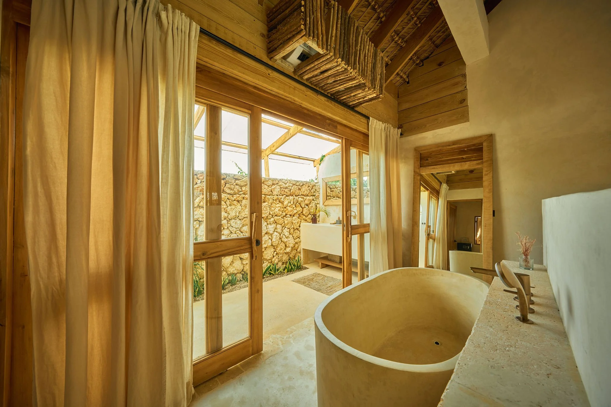 Vista de un baño con paredes de madera y una bañera de piedra, con puertas de cristal y cortinas beige que llevan a un patio con muro de piedra y jardín exterior.