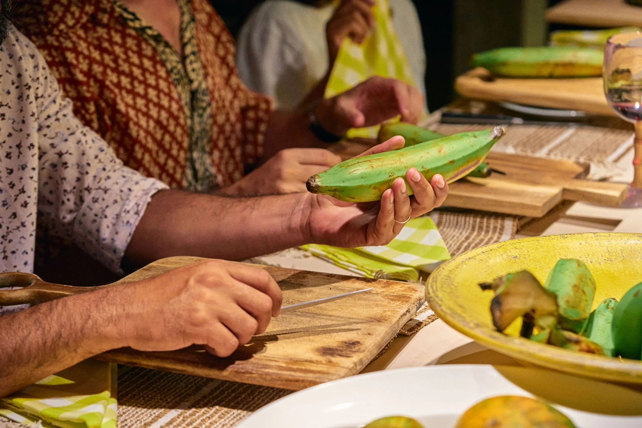 Varias personas pelando plátanos en una mesa de madera con manteles y plato amarillo, preparándolos para cocinar.