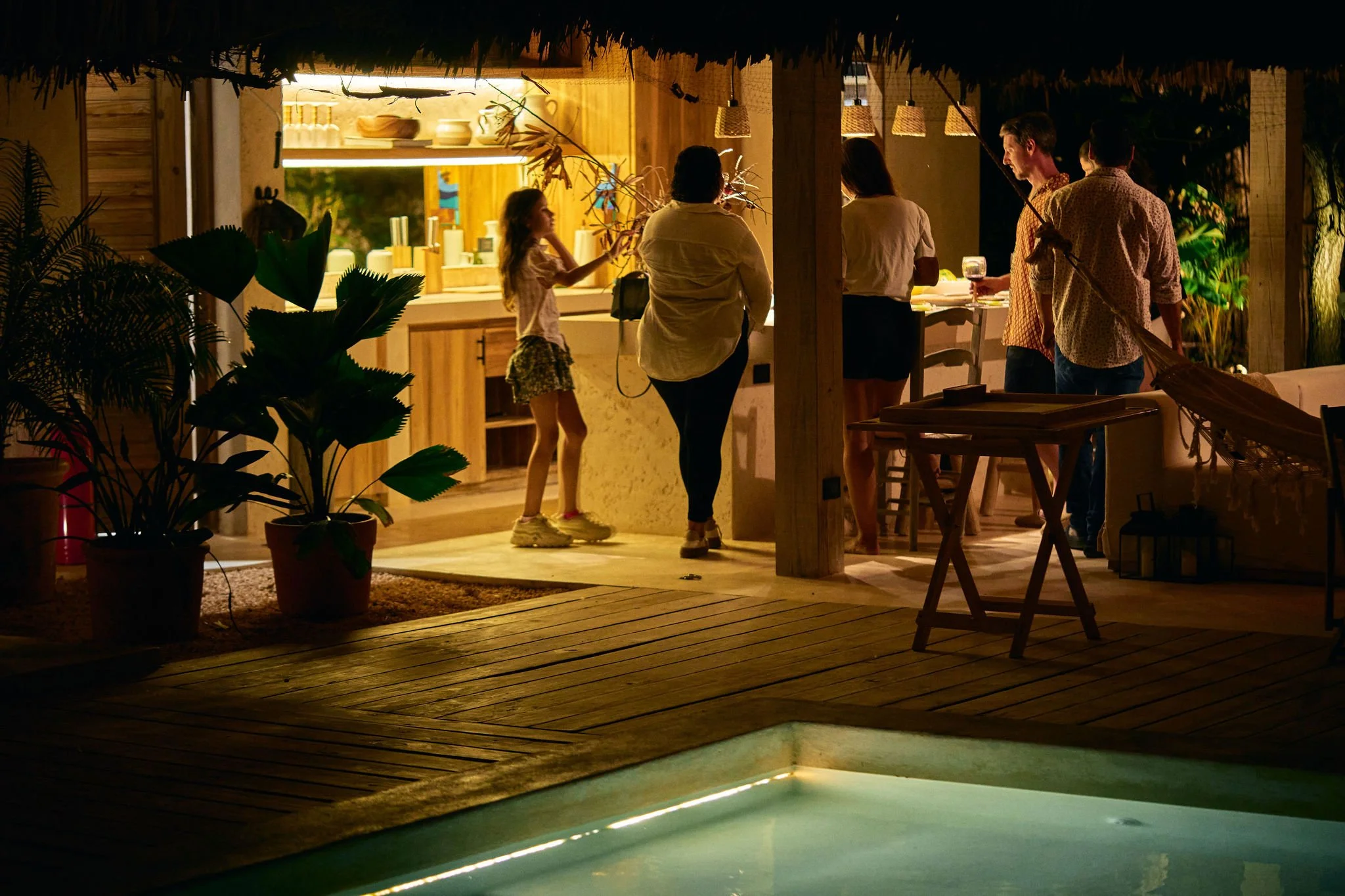 Grupo de personas en una terraza o área de piscina en una noche, conversando y disfrutando de bebidas, rodeados de plantas decorativas.