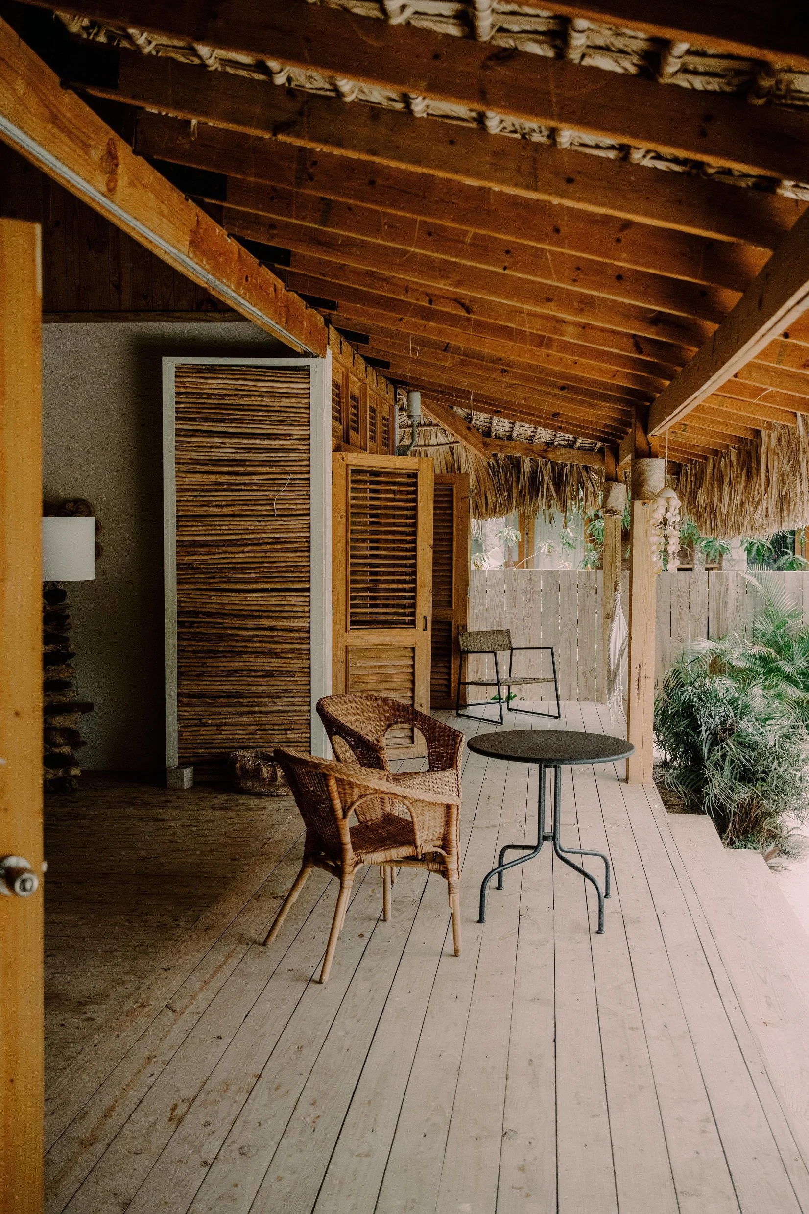 Una terraza de madera con sillas de mimbre, una mesa y plantas verdes en el fondo, con un techo de palmas.