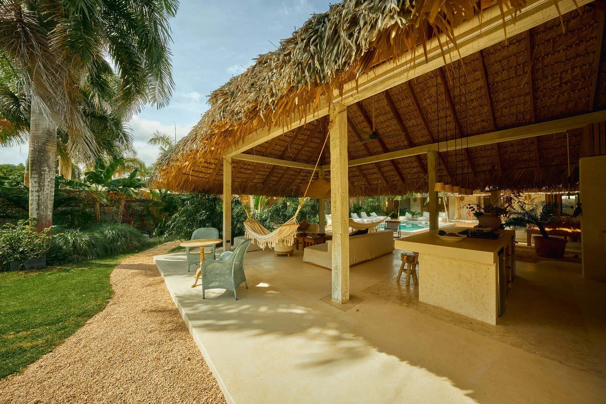 Ático con techo de paja, silla de mimbre, hamaca de cuerda, y vista a un jardín con árboles tropicales y piscina.